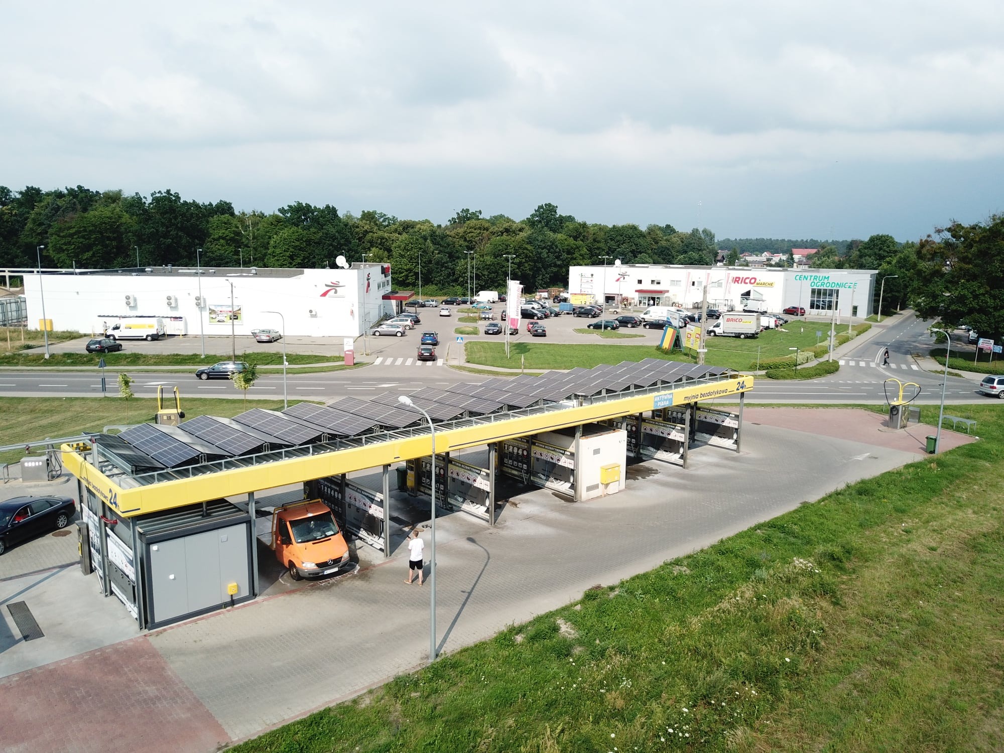 Widok z góry na myjnię bezdotykową z panelami słonecznymi na dachu, pomarańczowy bus w trakcie mycia, w tle budynki handlowe i drzewa.