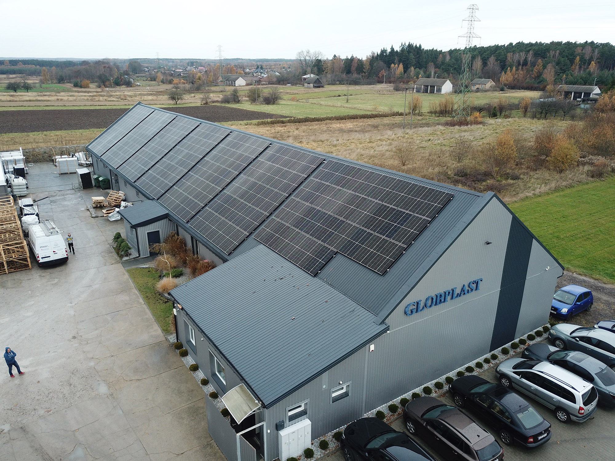 Widok z góry na szary budynek firmy Globplast z panelami słonecznymi na dachu, zaparkowanymi samochodami i osobą w czerwonych butach na betonowym placu.