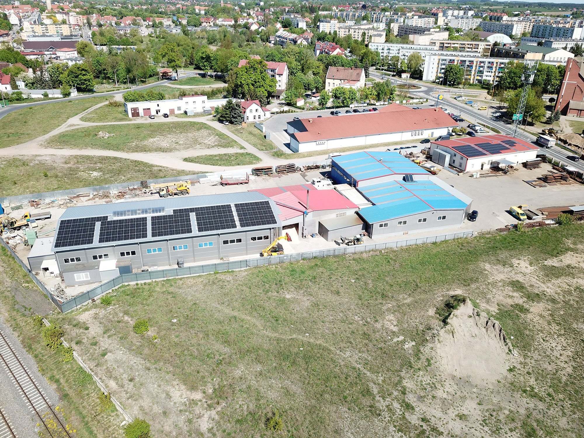 Widok z lotu ptaka na budynki przemysłowe z panelami słonecznymi na dachach, teren budowy z maszynami, w tle zabudowa miejska.