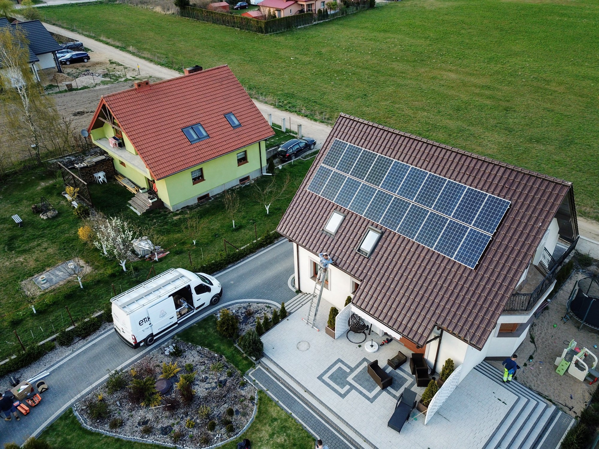 Widok z lotu ptaka na dom z panelami słonecznymi na dachu, instalator na drabinie, biały van firmy z otwartymi drzwiami i narzędziami, oraz zadbany ogród z kostką brukową.