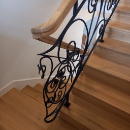 Promet - Drewniane schody z ozdobną, kutą, czarną balustradą. Poręcz drewniana, jasna. W tle widoczna osoba.