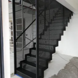 Czarne ażurowe schody z metalu, zabezpieczone siatką z drobnymi otworami, widoczne ślady prac wykończeniowych na posadzce.