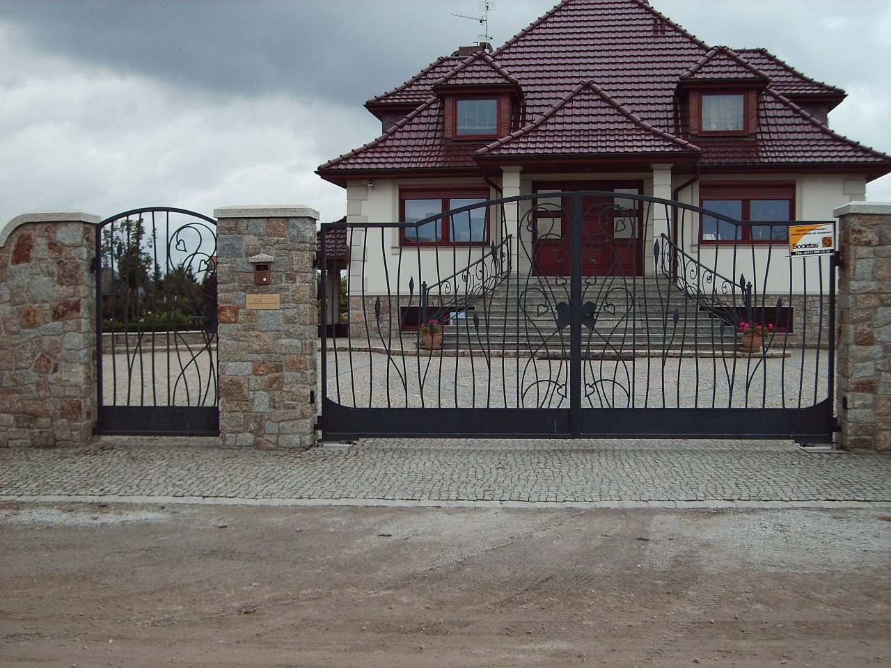 Czarna, metalowa brama wjazdowa z dekoracyjnymi elementami, osadzona pomiędzy kamiennymi słupkami na tle dwupiętrowego domu z czerwoną dachówką.