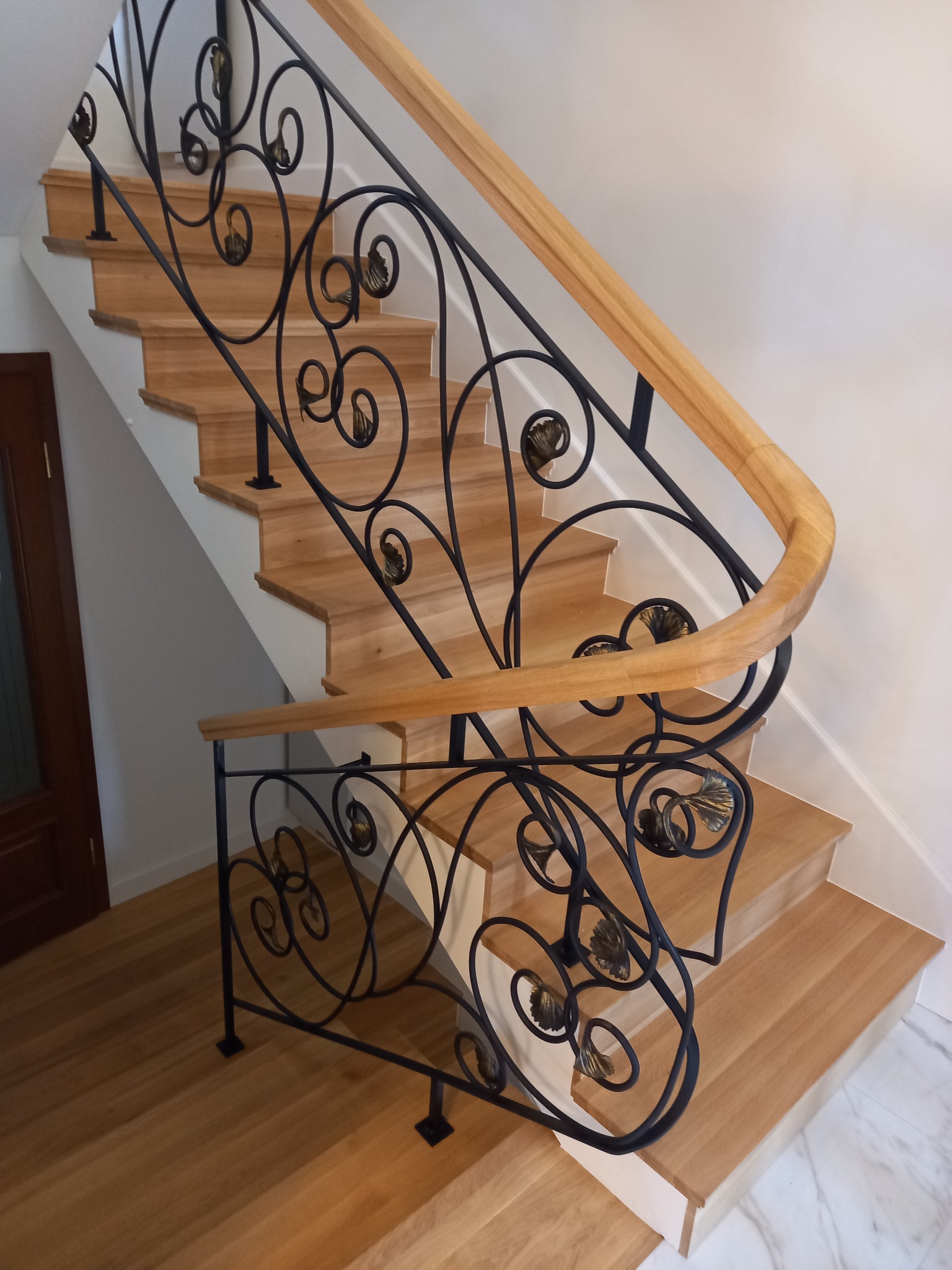 Drewniane schody z czarną, ozdobną balustradą z motywem liści i jasną, drewnianą poręczą, widoczne z góry.