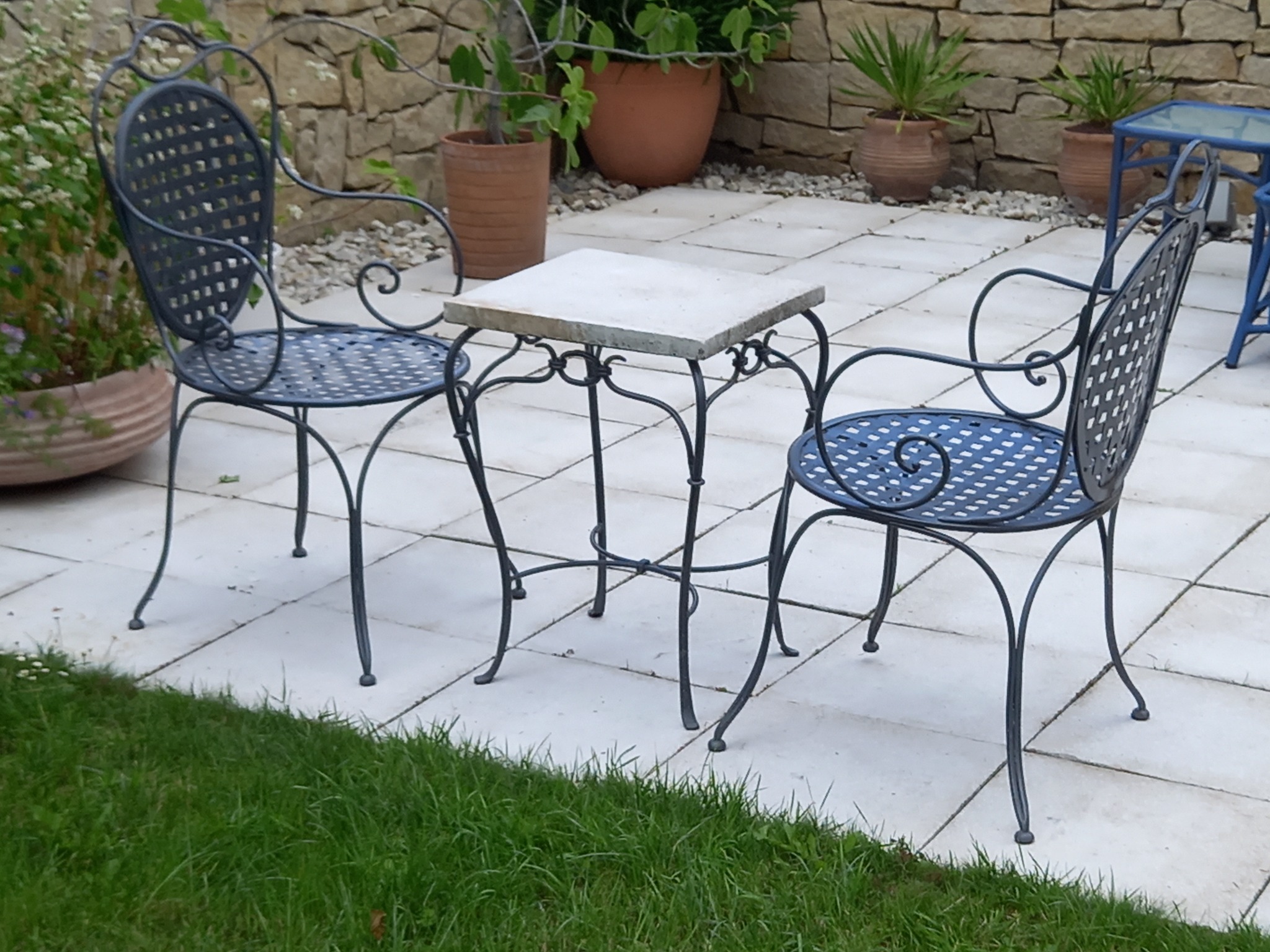 Dwa krzesła z metalową konstrukcją i ażurowym siedziskiem oraz stolik z kamiennym blatem, ustawione na jasnym brukowanym tarasie w ogrodzie, z widocznymi roślinami w donicach i kamiennym murem w tle.