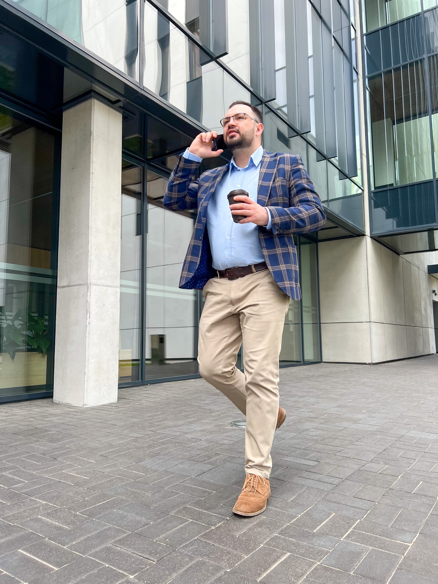 Mężczyzna w okularach i marynarce w kratę rozmawia przez telefon, trzymając kubek z kawą na tle nowoczesnego budynku biurowego z przeszkloną fasadą.
