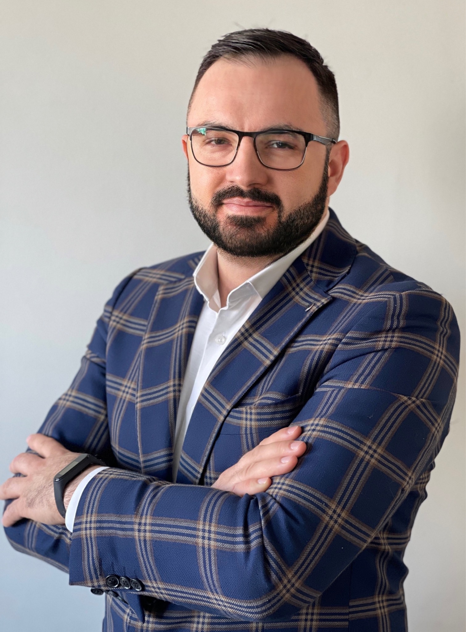 Portret mężczyzny w okularach i niebieskiej marynarce w kratę, z rękami skrzyżowanymi na piersiach, na jasnym tle.