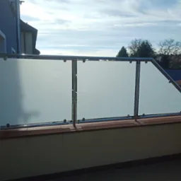 Balkon z balustradą ze stali nierdzewnej i szkła matowego, widok na zabudowania i niebo z chmurami.