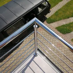 Błyszcząca, metalowa balustrada z wypełnieniem z cienkich prętów, widok z góry, w tle czarny bus zaparkowany na kostce brukowej.