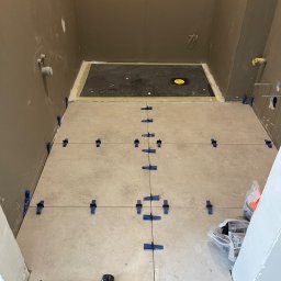 TMT projects - Układanie jasnych płytek podłogowych w łazience, widoczne krzyżyki dystansowe, sznurek wyznaczający linie i gumowy młotek przy progu.