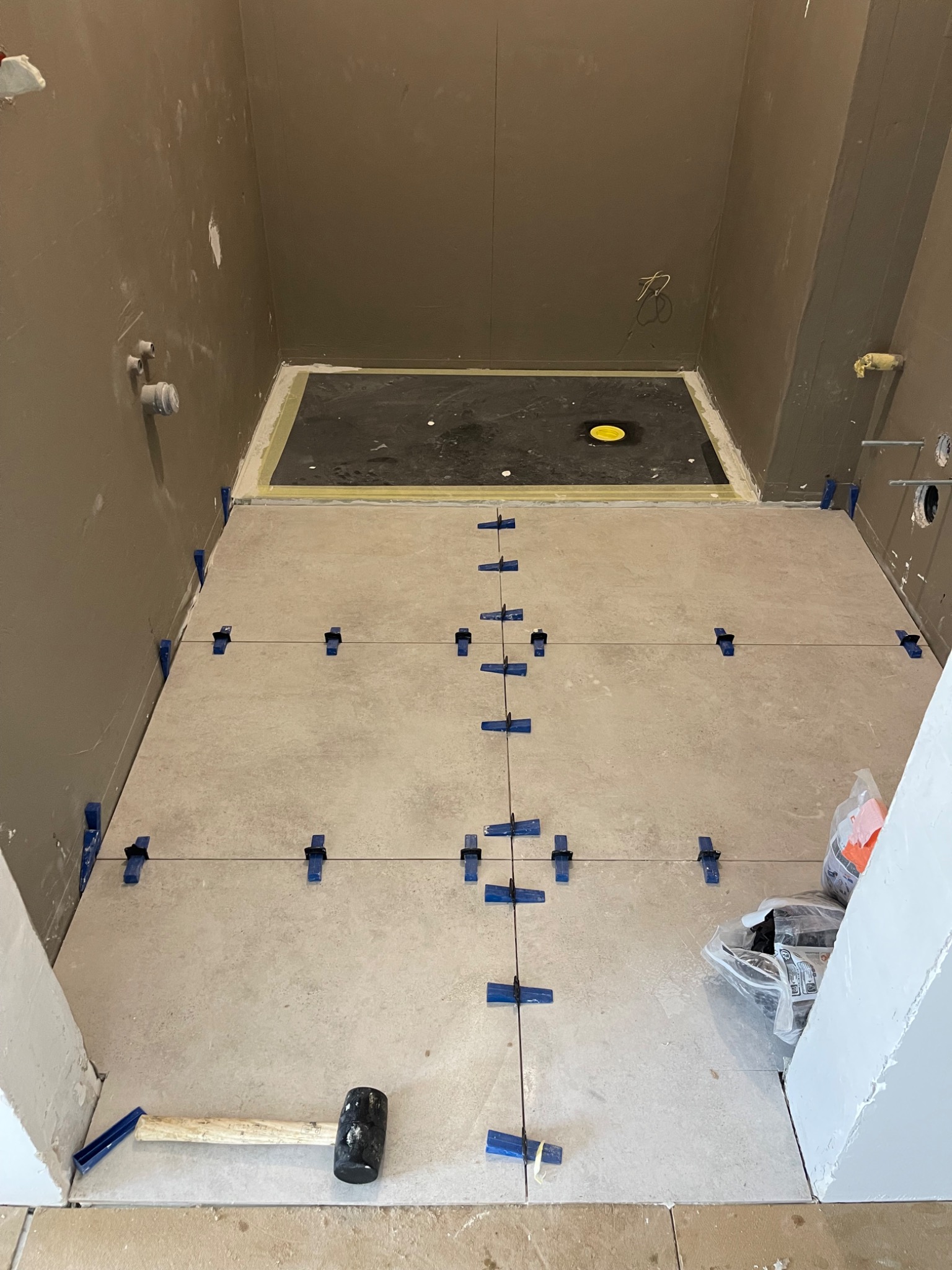 Układanie jasnych płytek podłogowych w łazience, widoczne krzyżyki dystansowe, sznurek wyznaczający linie i gumowy młotek przy progu.