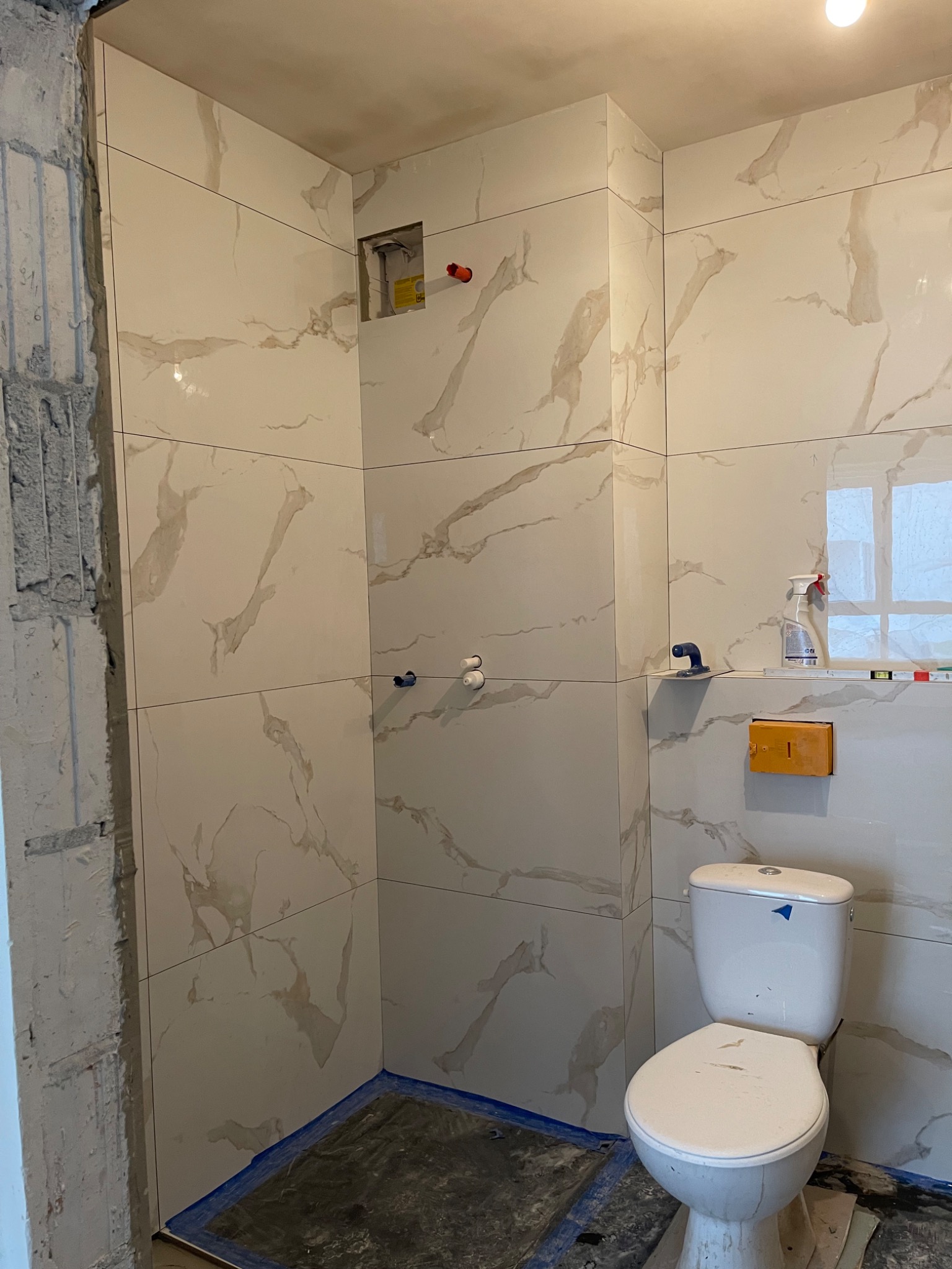 Montaż płytek imitujących marmur w łazience, widoczny fragment surowej ściany, toaleta z niebieską naklejką, przygotowane otwory na armaturę i akcesoria łazienkowe.