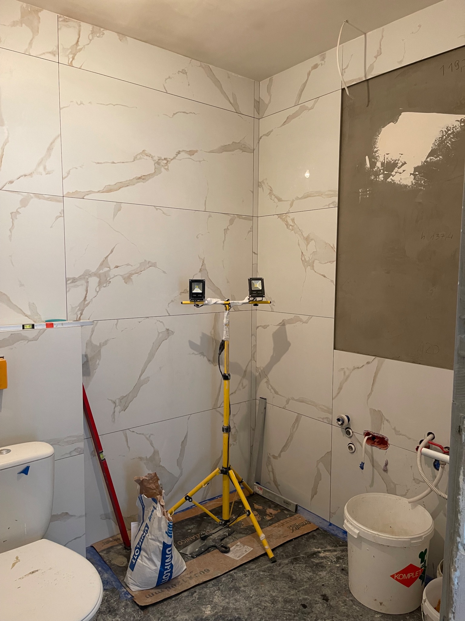 Łazienka w trakcie układania płytek imitujących marmur, widoczne narzędzia, poziomica, worki z zaprawą, lampy budowlane na statywie i fragment ściany przygotowany pod płytki.