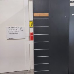 Mg Tramo Sp z o.o. - Ciemnoszara szafa z drewnianym akcentem w górnej części, widoczna w surowym wnętrzu hali produkcyjnej obok tablicy z informacją o zakazie wstępu dla montażystów z powodu COVID-19.