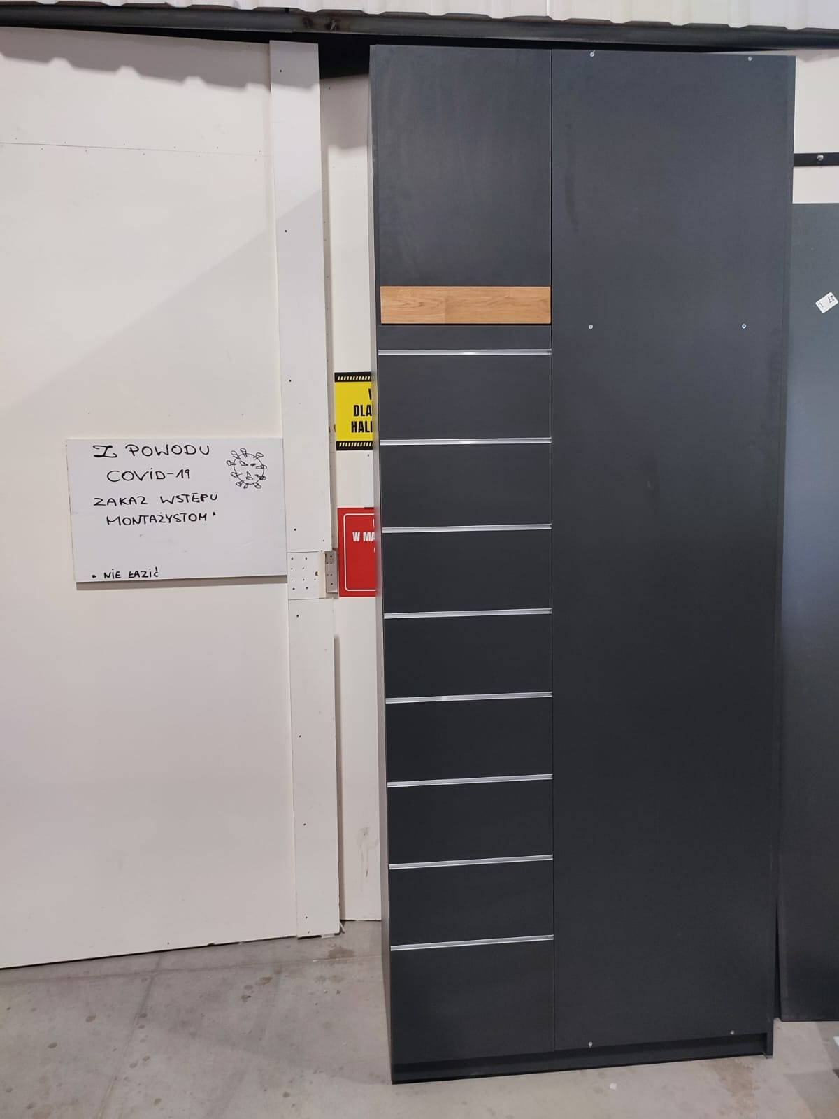 Ciemnoszara szafa z drewnianym akcentem w górnej części, widoczna w surowym wnętrzu hali produkcyjnej obok tablicy z informacją o zakazie wstępu dla montażystów z powodu COVID-19.
