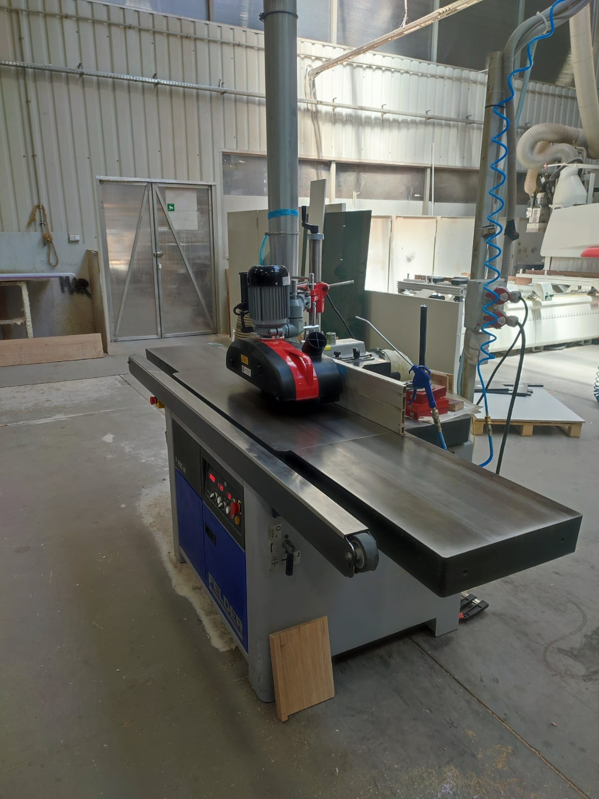 Profesjonalna frezarka Felder F 500 M w warsztacie, z widocznym panelem sterowania, elementami zabezpieczającymi i fragmentem obrabianego drewna u podstawy. W tle widoczne inne maszyny i elementy...