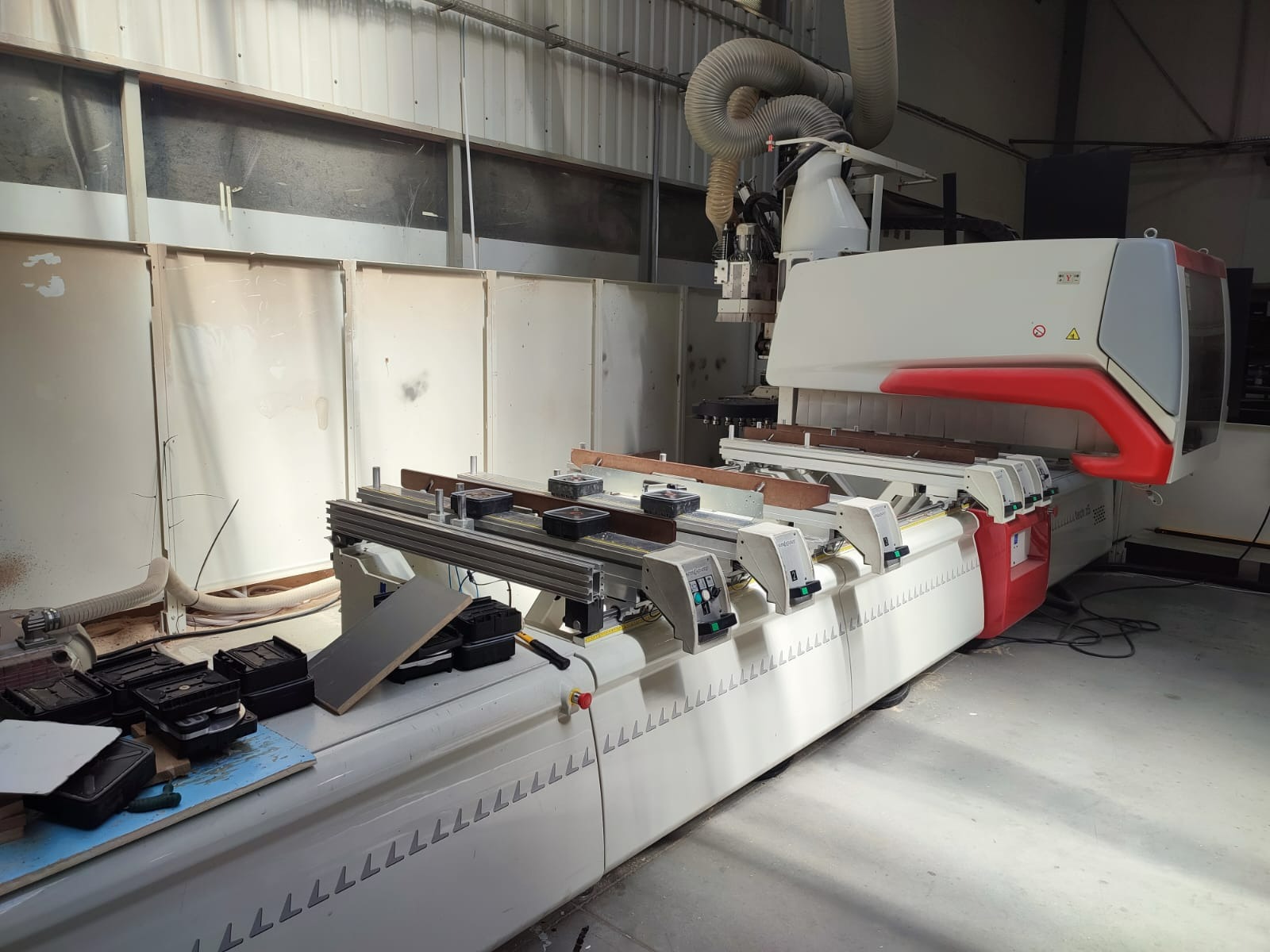 Profesjonalna maszyna CNC do obróbki drewna w warsztacie, widoczne elementy mocujące i system odciągu pyłu, oświetlenie halogenowe. Elementy drewniane zamocowane na stole roboczym.