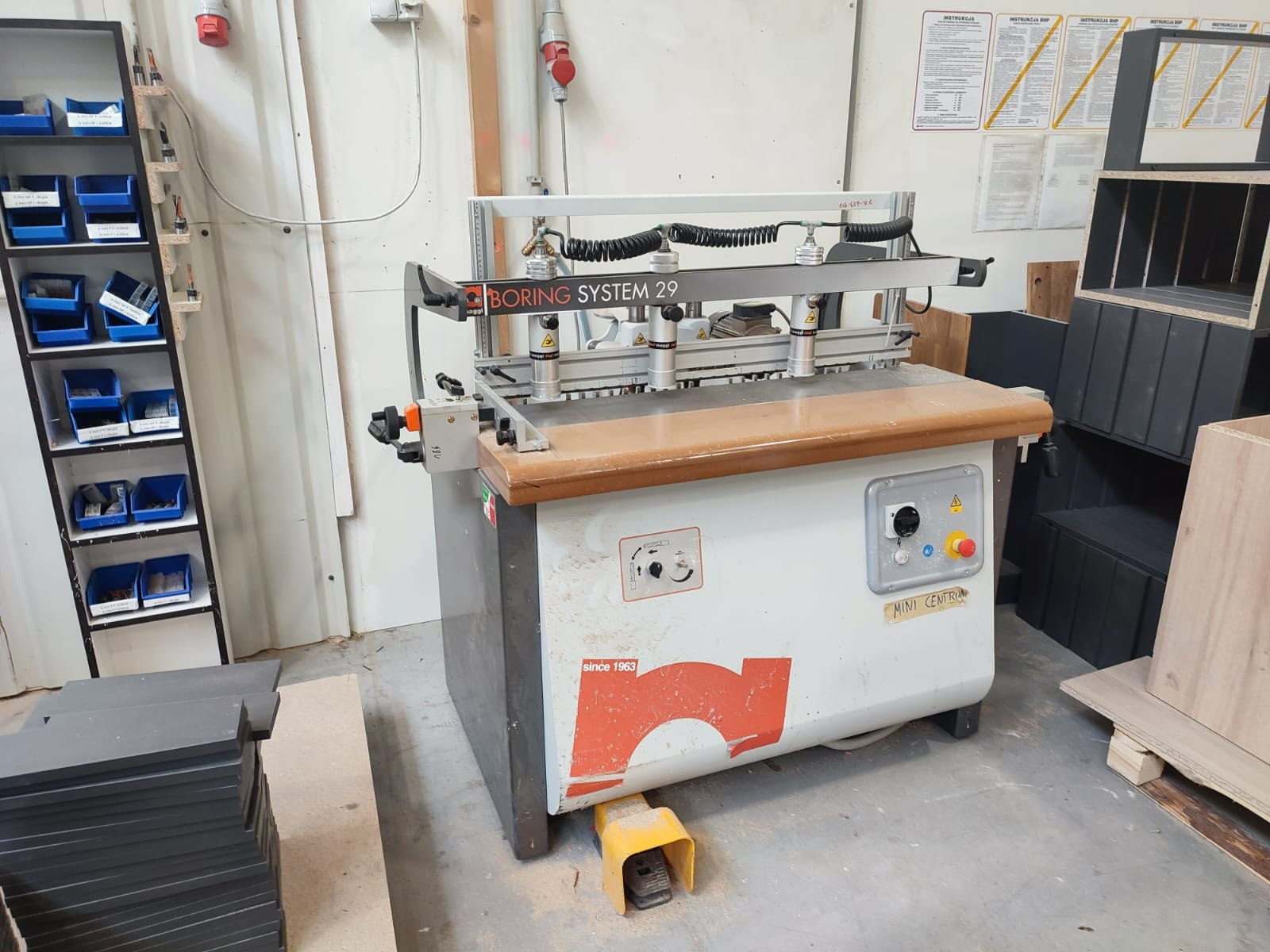 Wiertarka wielowrzecionowa BORING SYSTEM 29 w warsztacie, obok regał z pojemnikami na narzędzia i stosem płyt meblowych.