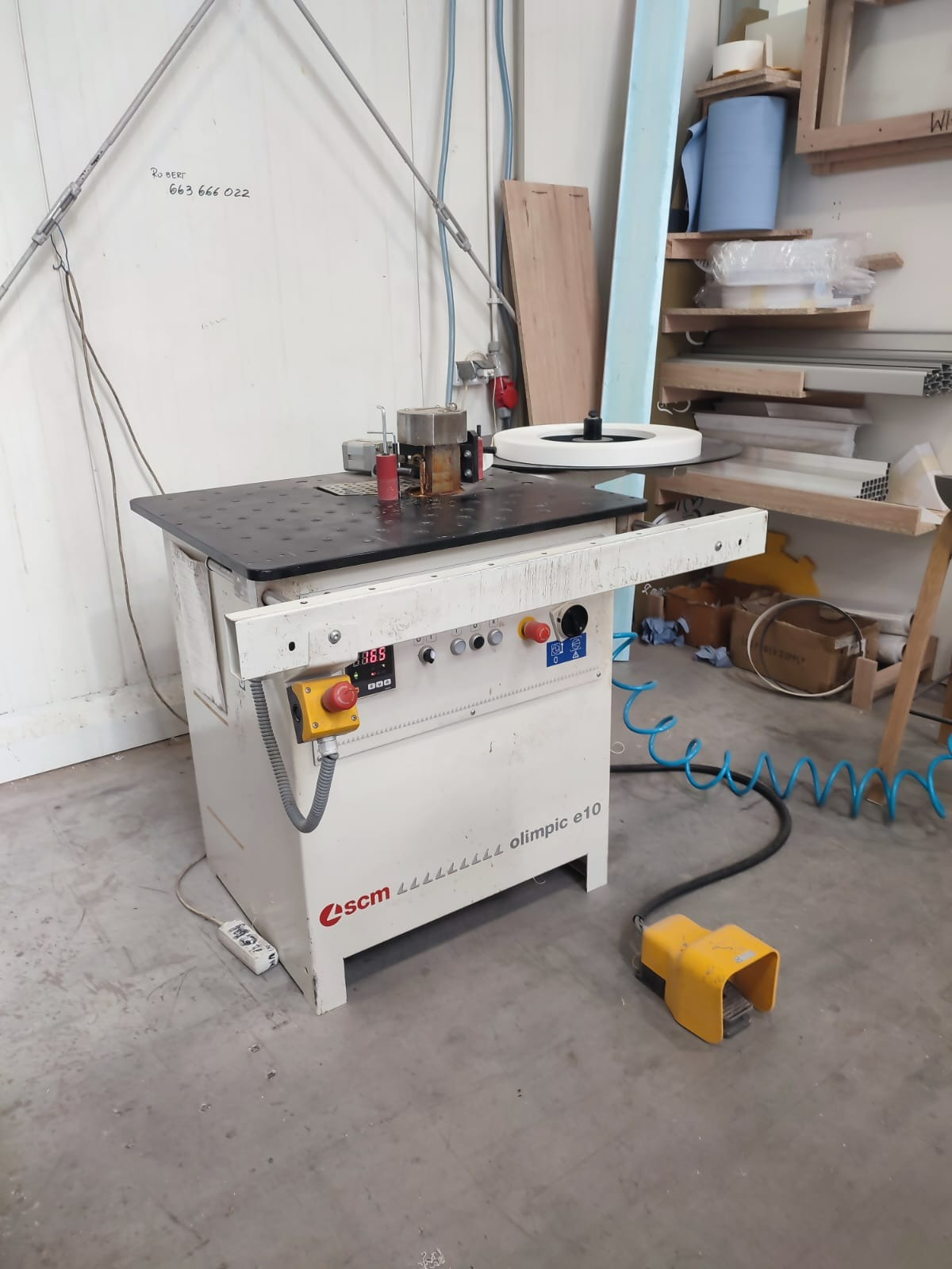 Frezarka krawędziowa SCM Olimpic e10 w warsztacie, widoczne narzędzia, przewody, pedał nożny, oraz półki z materiałami w tle.