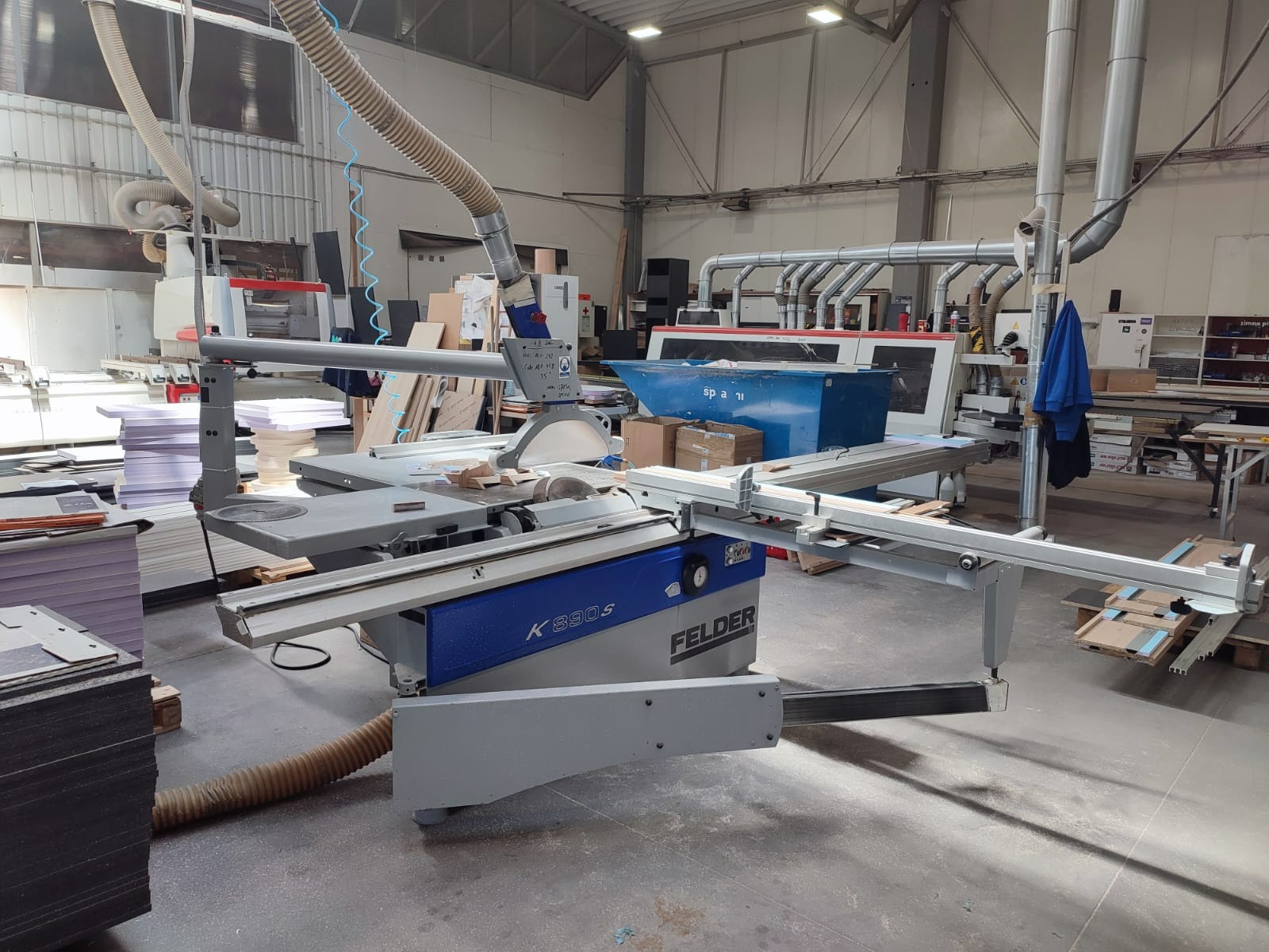 Piła formatowa Felder K 390 S w warsztacie, z widocznymi elementami odciągu trocin i stosem płyt meblowych w tle.
