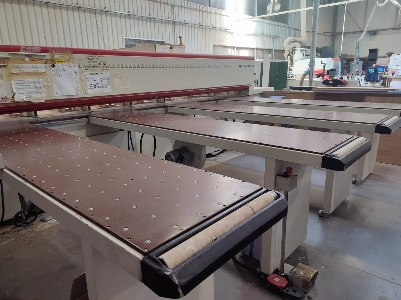 Profesjonalna piła panelowa Sigma Prima w warsztacie stolarskim, z widocznymi blatami roboczymi z otworami i rolkami dociskowymi.