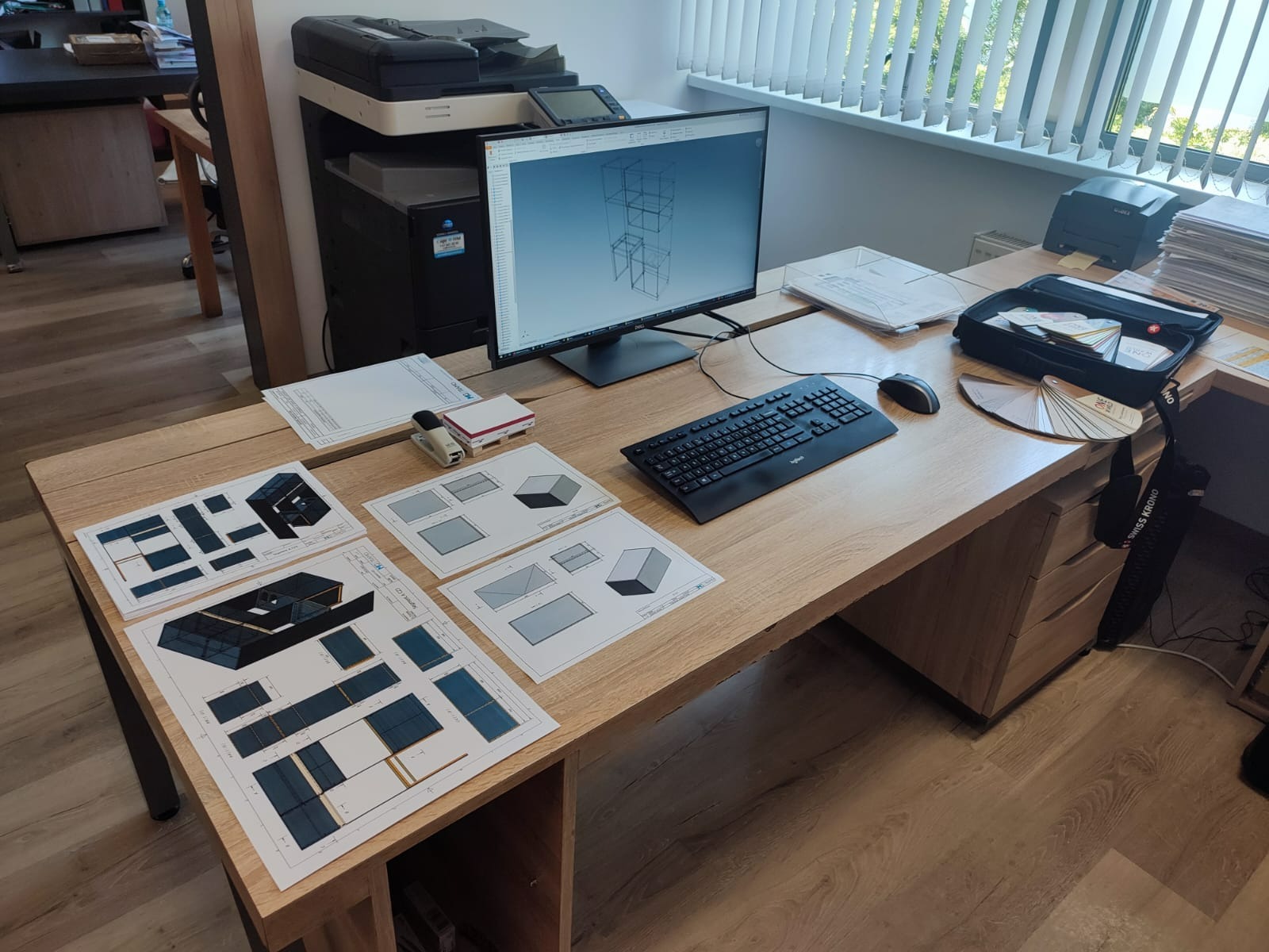 Biurko z komputerem wyświetlającym model 3D konstrukcji meblowej oraz rozłożonymi planami technicznymi, próbnikami kolorów i akcesoriami biurowymi, sugerujące proces projektowania.