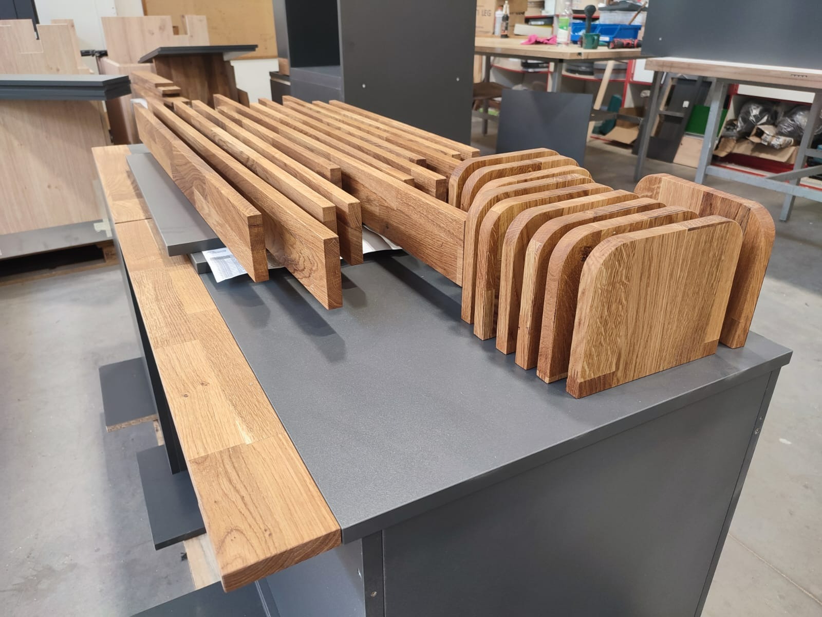 Przygotowane elementy drewniane, prawdopodobnie do mebli, ułożone na szarym blacie w warsztacie stolarskim. Widoczne proste listwy i zaokrąglone płyty z wyraźnym rysunkiem drewna dębowego.