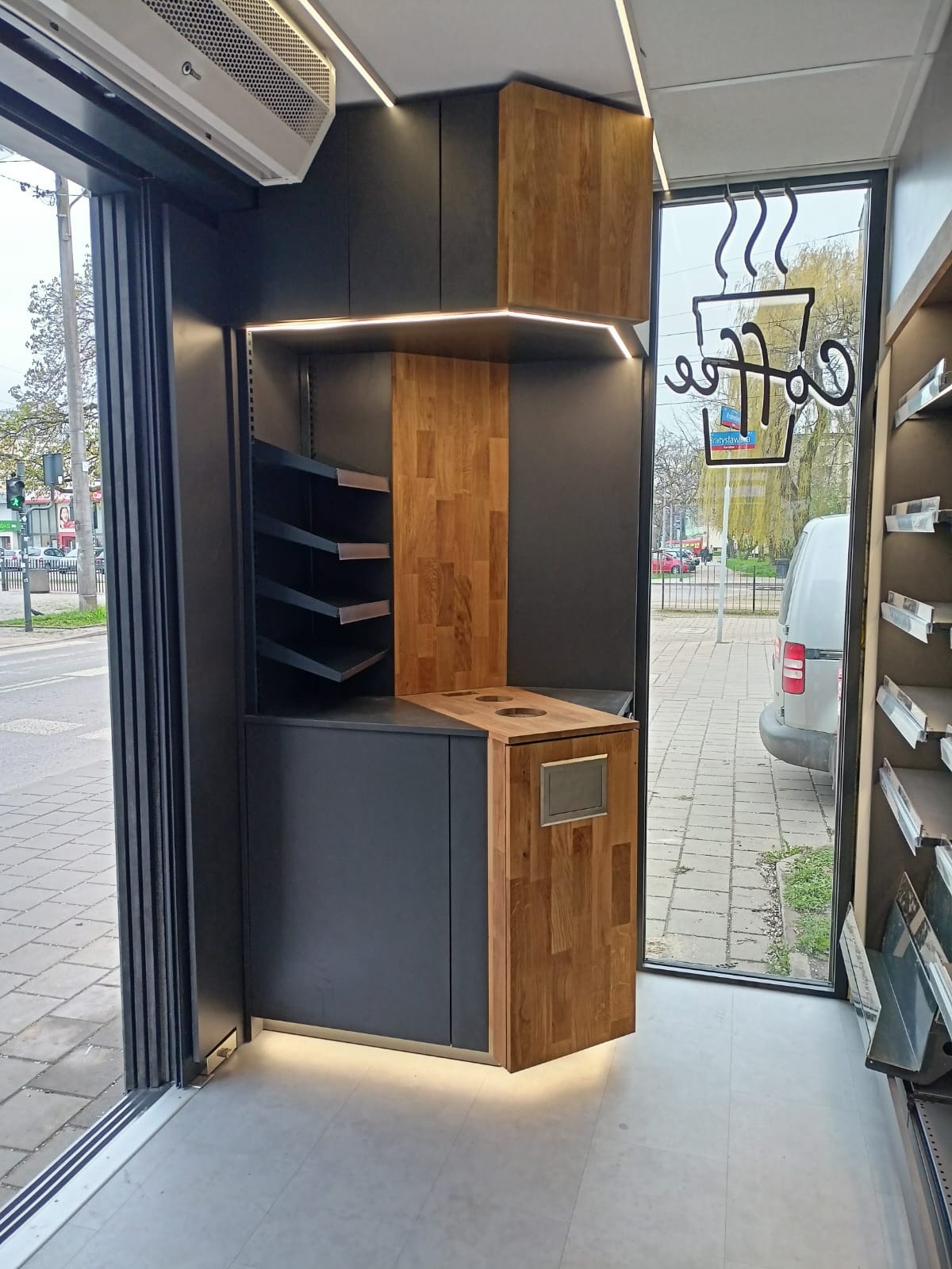 Czarno-drewniana zabudowa meblowa z oświetleniem LED w lokalu usługowym, widoczny fragment ulicy przez okno z naklejką 'Coffee'.