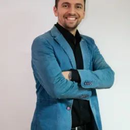 Uśmiechnięty mężczyzna w dżinsowej marynarce i czarnej koszuli, stojący na tle jasnej ściany z założonymi rękami i zegarkiem na nadgarstku.