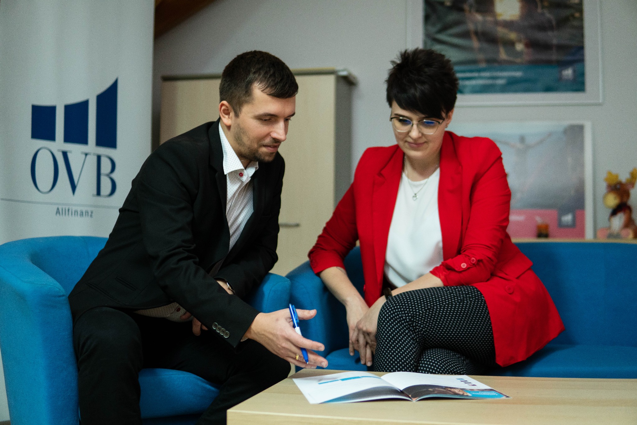 Mężczyzna w garniturze i kobieta w czerwonej marynarce omawiają dokumenty przy stole, w tle logo OVB Allfinanz na banerze.