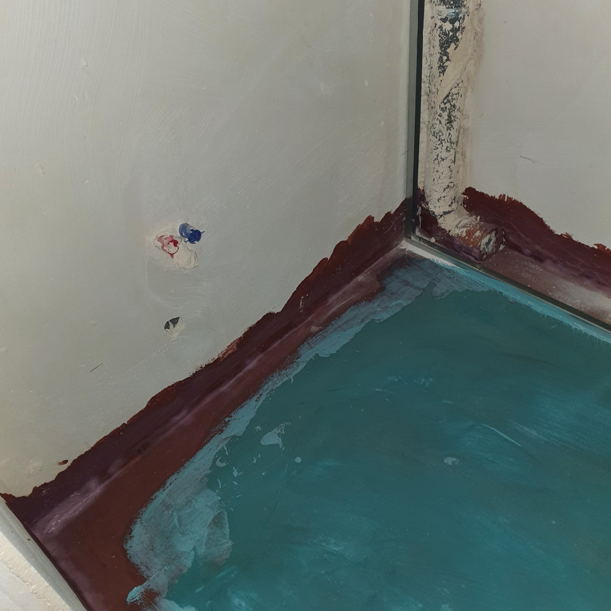 Hydroizolacja podłoża w rogu łazienki z widocznymi śladami po pędzlu i zabezpieczeniem na ścianach, przygotowanie do dalszych prac wykończeniowych.