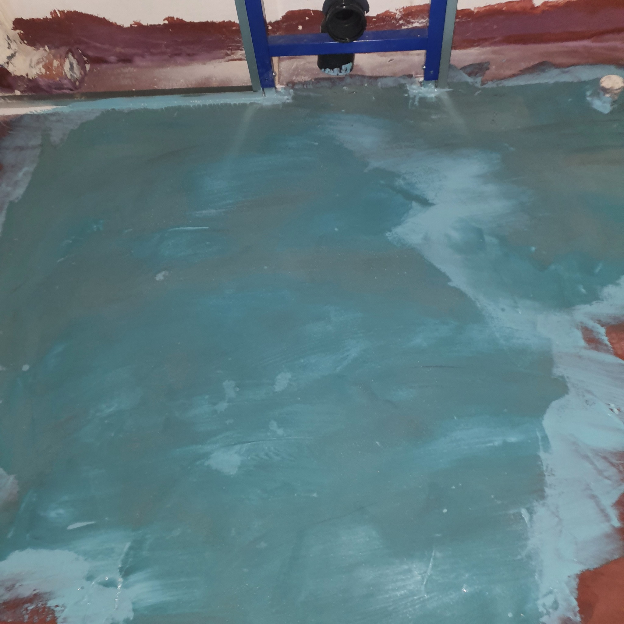 Nierównomiernie rozprowadzona, turkusowa warstwa wylewki samopoziomującej na podłodze łazienki, widoczne ślady po szpachli na ścianach i stelaż podtynkowy WC.