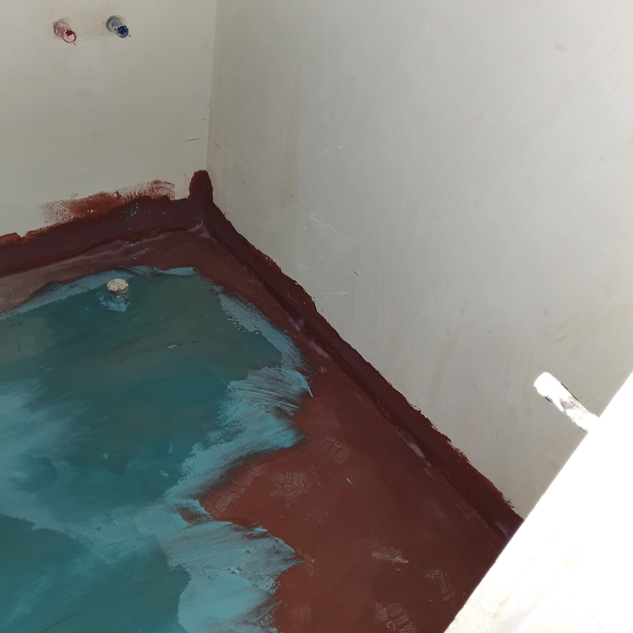 Przygotowanie podłoża w łazience w Wałczu: brązowa hydroizolacja na ścianach i podłodze, częściowo pokryta turkusową warstwą, widoczne rury instalacji wodnej.