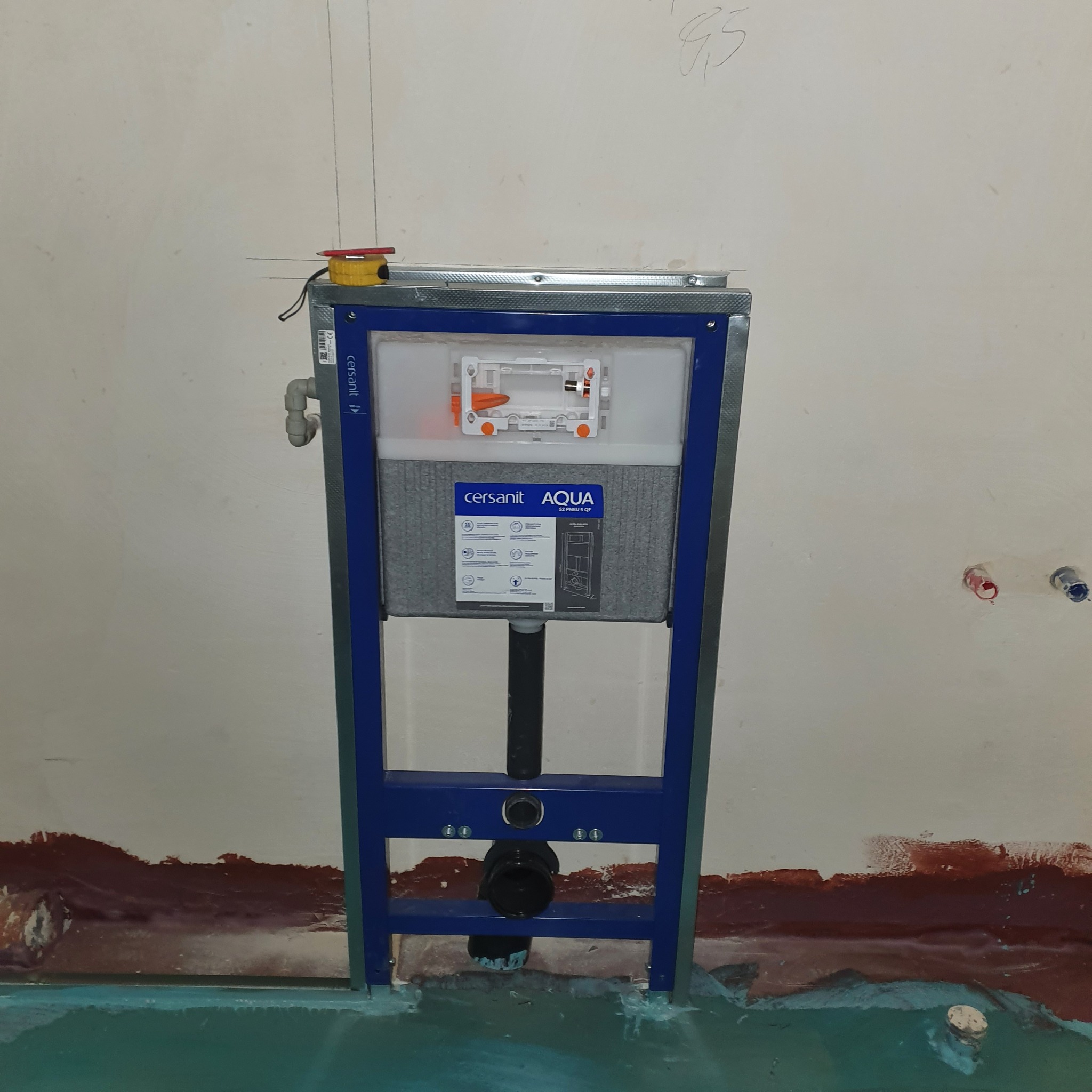 Stelaż podtynkowy WC Cersanit Aqua z widocznymi przyłączami wodnymi i odpływem kanalizacyjnym, zamontowany na tle surowej ściany z zaznaczonymi pionami i pozostałościami po pracach malarskich.