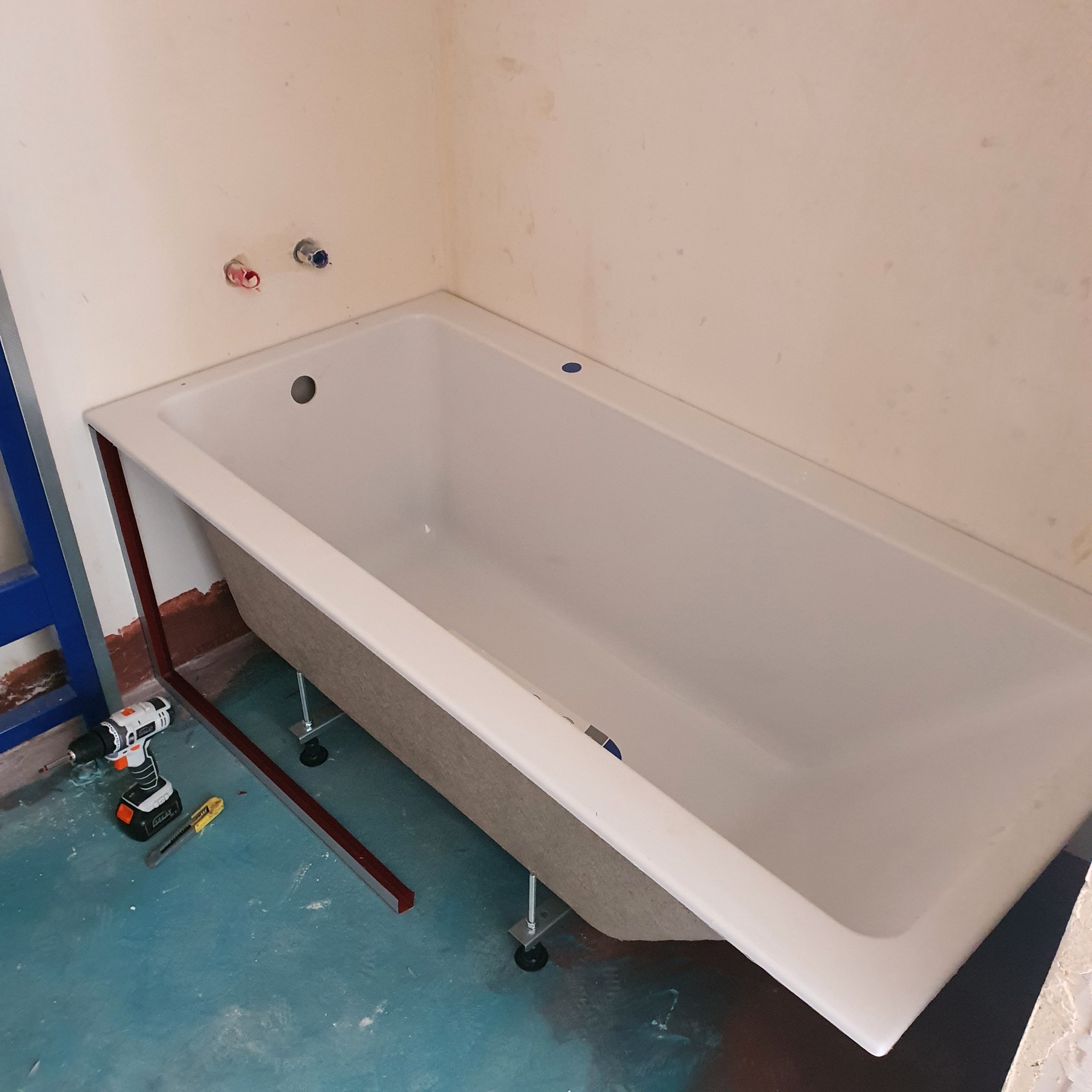 Biała wanna na regulowanych nóżkach w trakcie montażu w surowej łazience z widocznymi instalacjami wodnymi, wiertarką i nożykiem na podłodze.