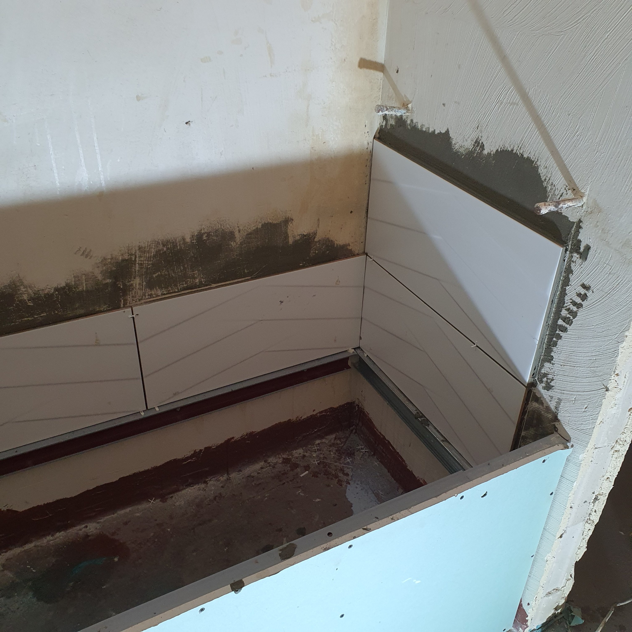 Niski kąt widzenia na częściowo wykończoną wnękę z białymi płytkami ułożonymi we wzór jodełki, widoczne metalowe profile i surowa ściana w tle.