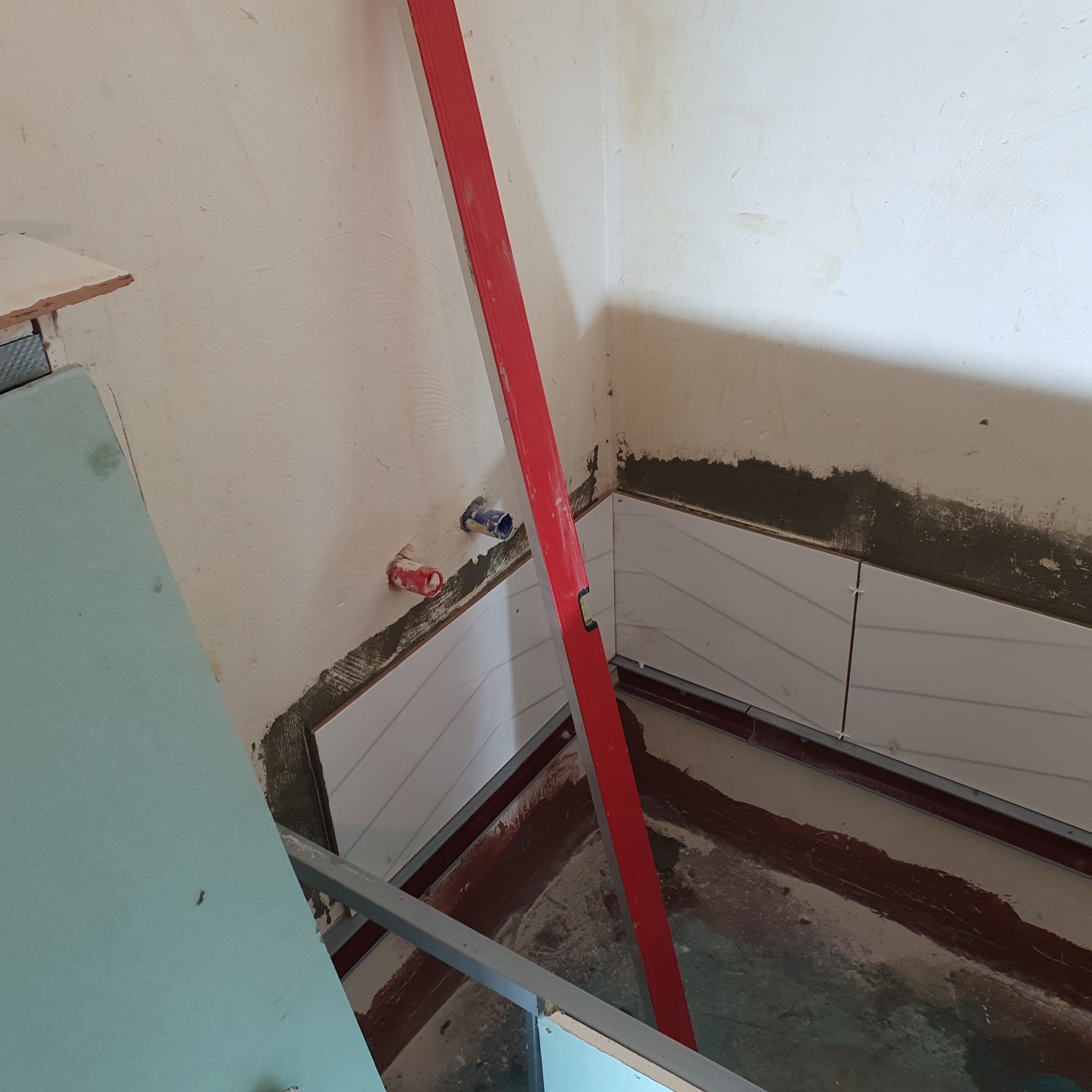 W trakcie układania białych, poziomych płytek w łazience, widoczny poziomica w kolorze czerwonym, fragment ściany z instalacją wodną i płyta karton gips na metalowym stelażu.