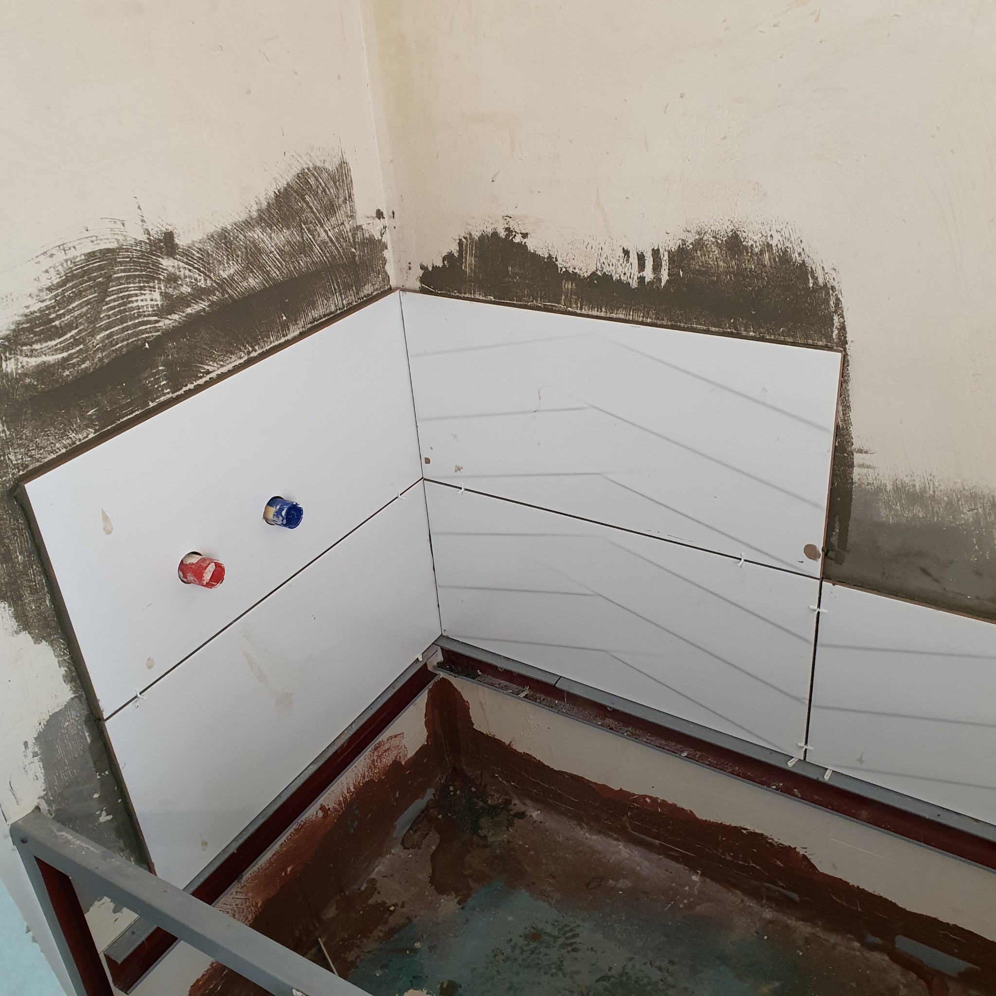 Kąt łazienki w trakcie układania białych, podłużnych płytek z delikatnym wzorem jodełki; widoczne wystające rury z instalacji wodnej, niezabezpieczone ściany z resztkami kleju i plastikowe krzyżyki...