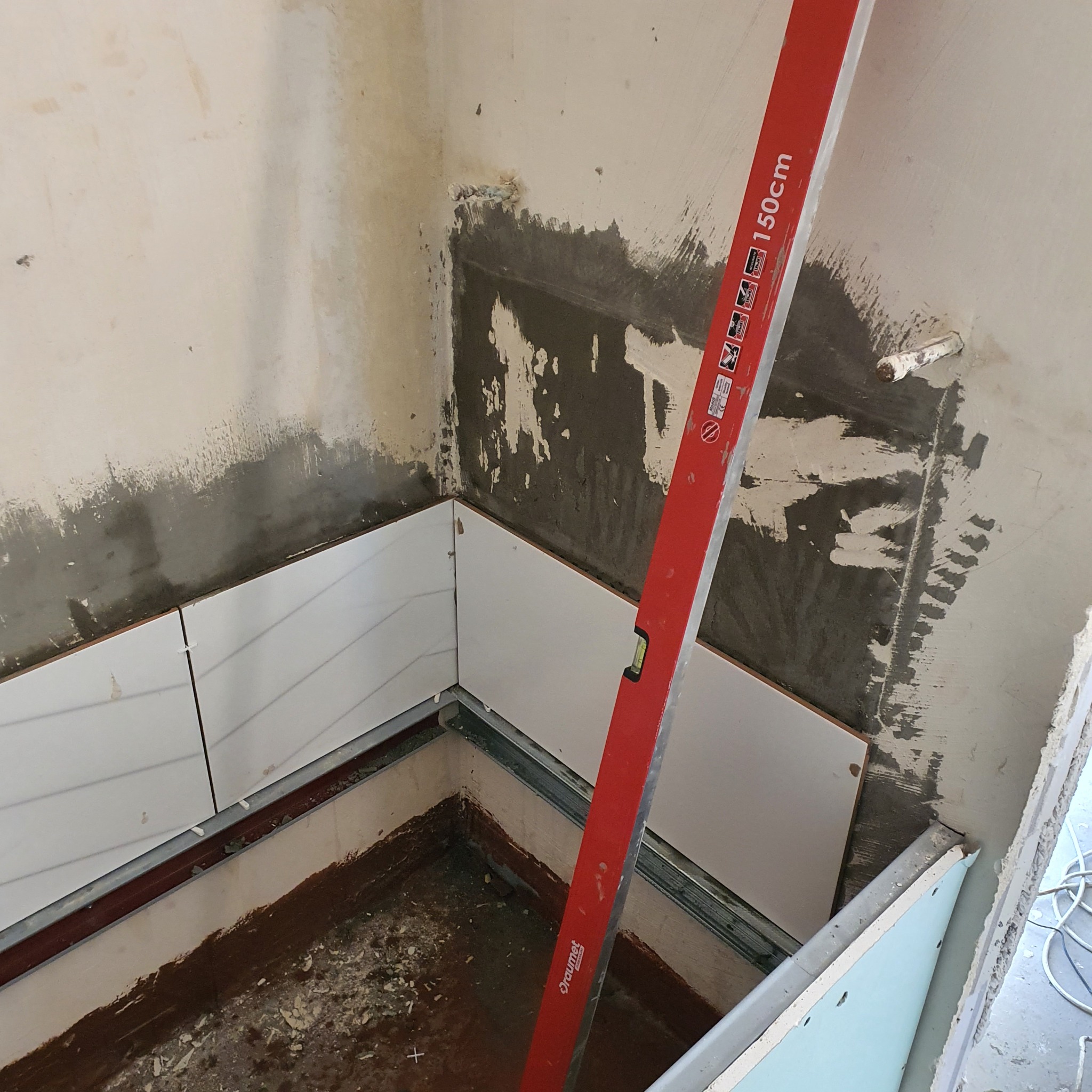 Fragment łazienki w trakcie prac wykończeniowych: białe płytki ułożone na dolnej części ściany, czerwona poziomica oparta o ścianę, widoczne ślady zaprawy i niedokończone prace.