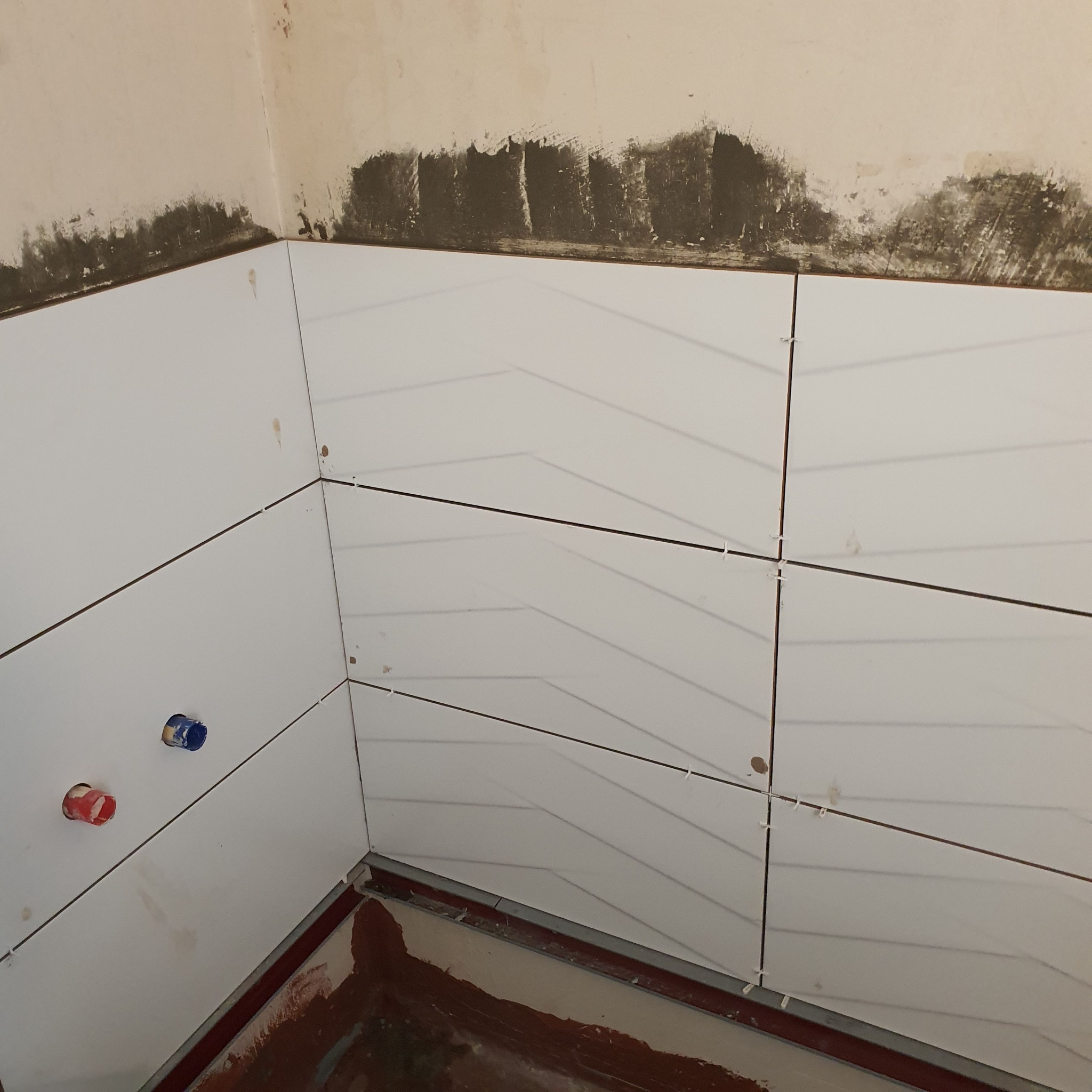 Białe, prostokątne płytki ułożone w jodełkę na ścianie w trakcie prac wykończeniowych w łazience, widoczne otwory na rury z instalacji wodnej oraz plastikowe krzyżyki dystansowe między płytkami.