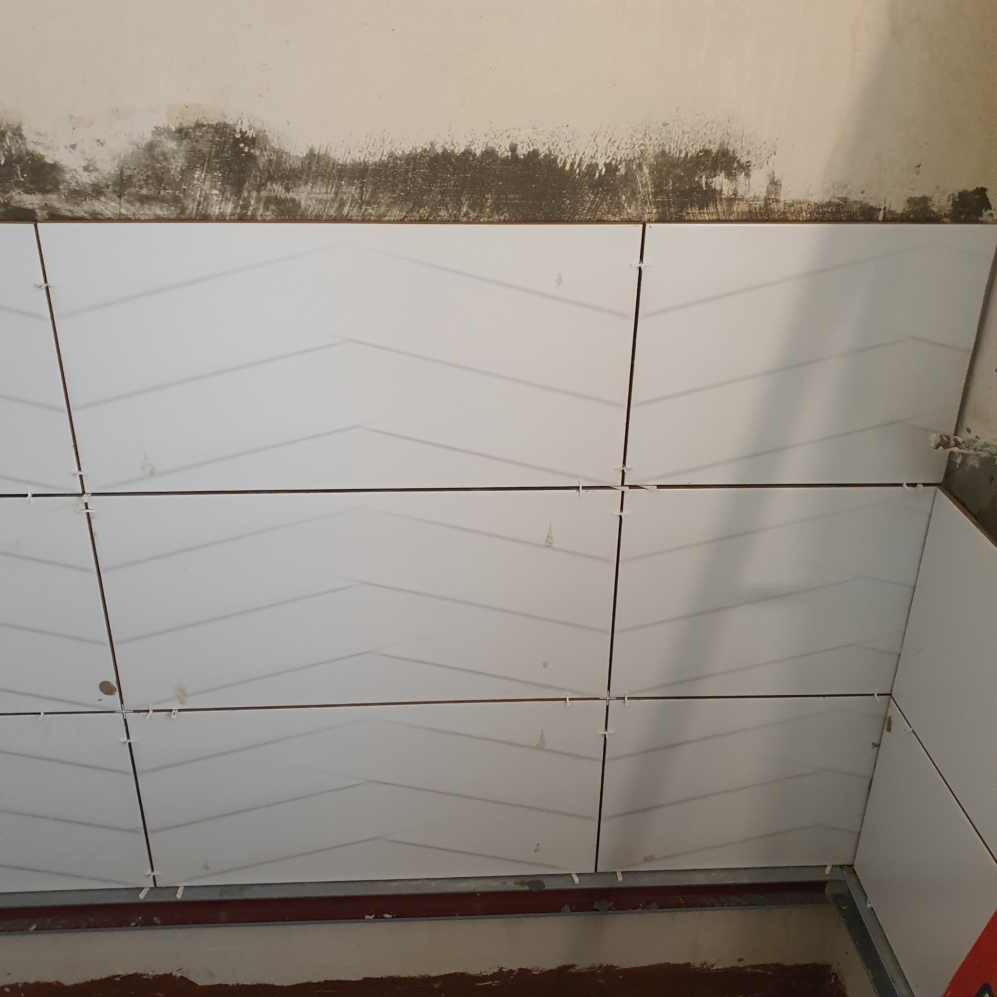 Białe płytki ceramiczne z delikatnym wzorem ułożone na ścianie w trakcie remontu łazienki, widoczne krzyżyki dystansowe i surowy stan ściany powyżej.