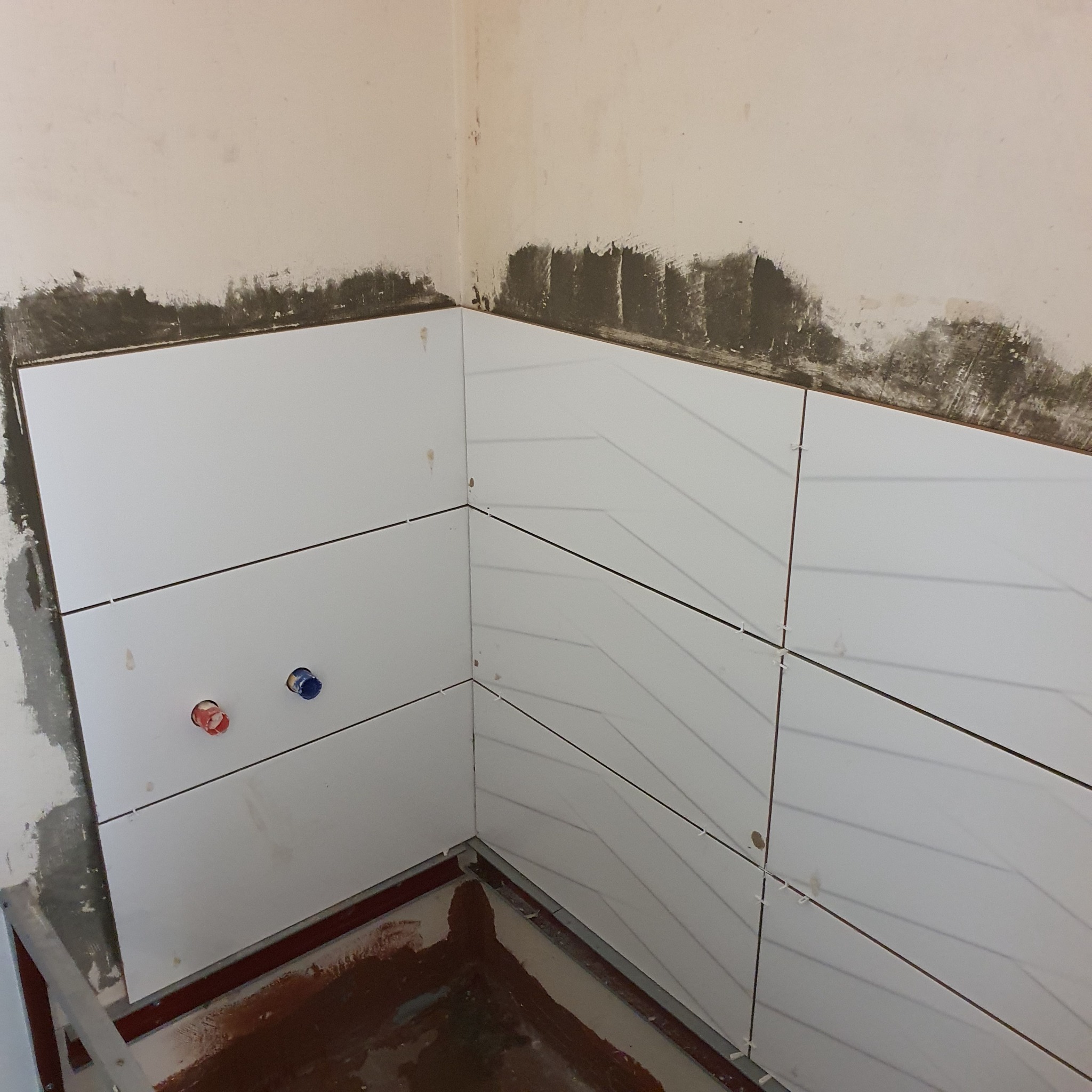 Układanie białych płytek w narożniku łazienki, widoczne krzyżyki dystansowe, wystające rury instalacji wodnej, ślady zaprawy na ścianie.