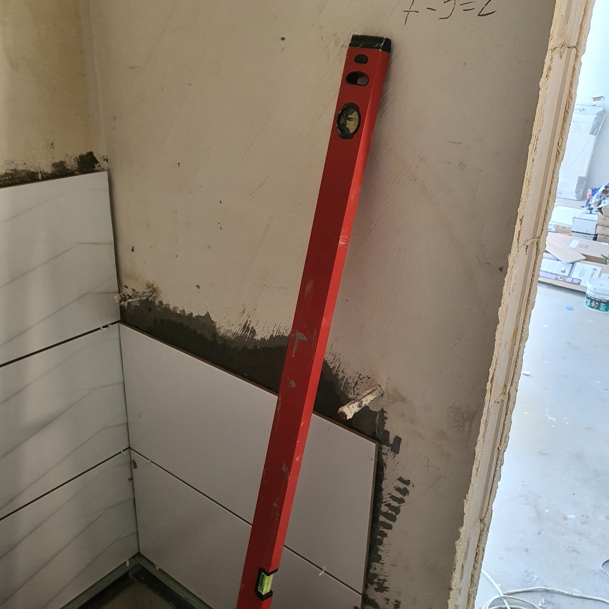 Układanie białych, prostokątnych płytek na ścianie w rogu pomieszczenia, widoczna poziomica oparta o ścianę, surowy stan wykończenia.