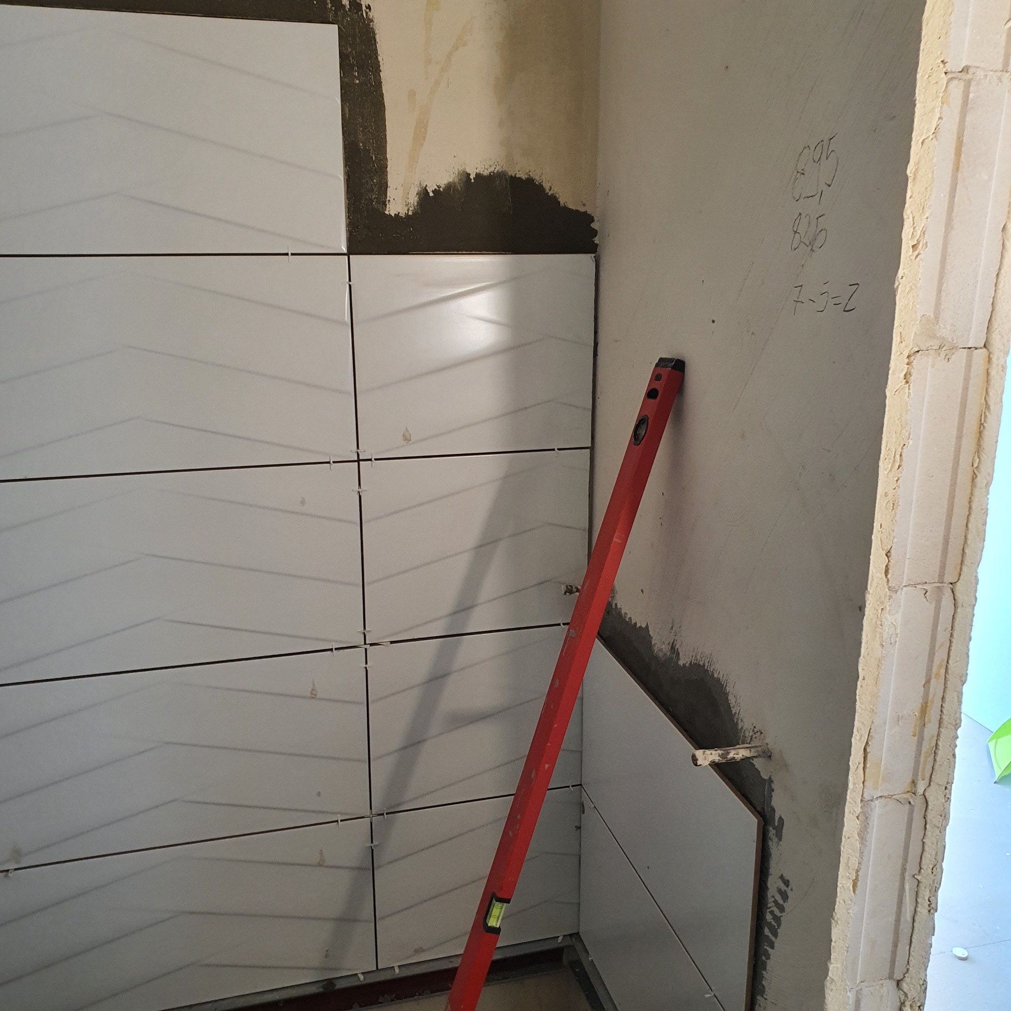 Układanie białych, podłużnych płytek z delikatnym wzorem na ścianie w rogu pomieszczenia. Widoczny czerwony poziom, szare ślady zaprawy, fugi i notatki na ścianie obok.