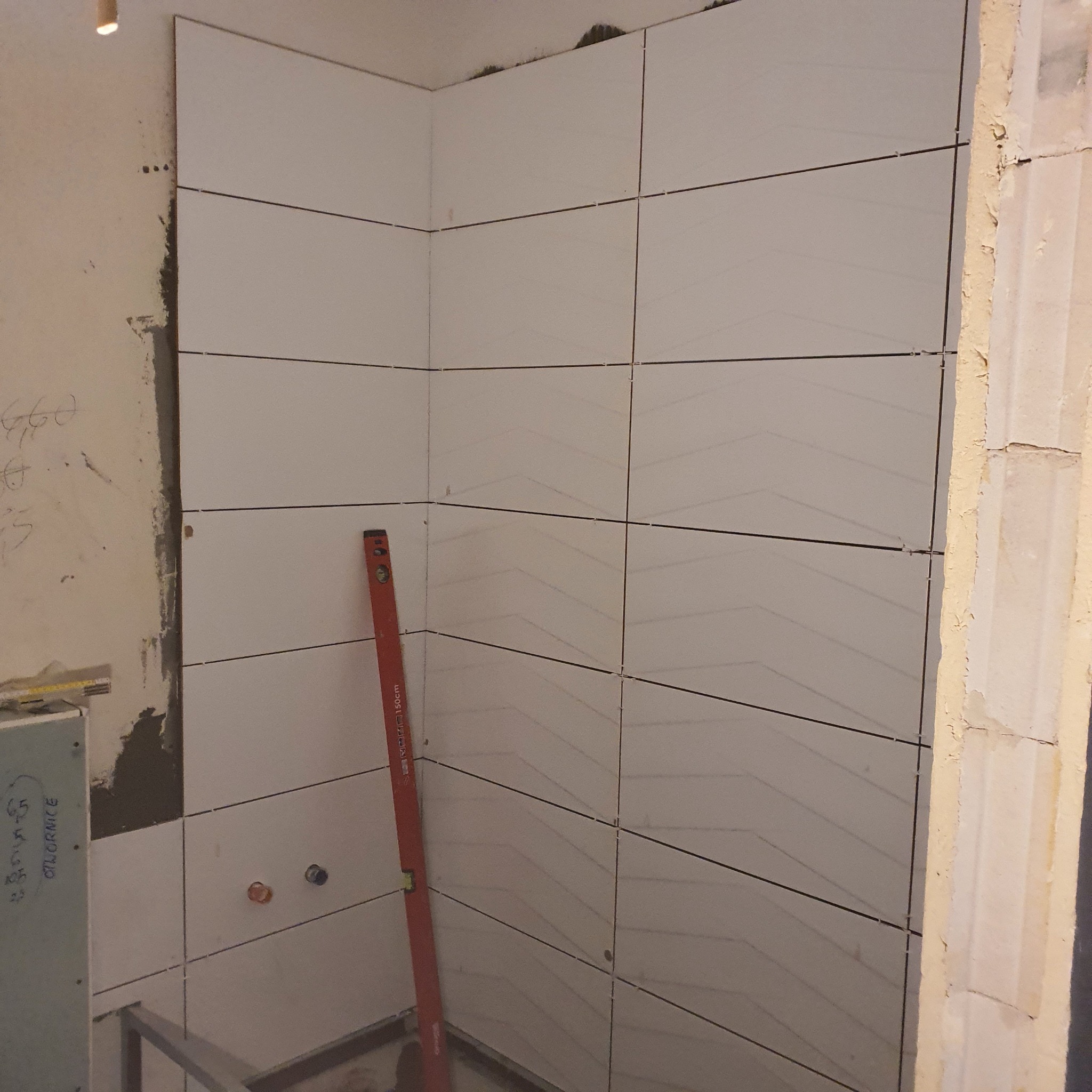 Świeżo ułożone, poziome, białe płytki w rogu łazienki, z widocznymi krzyżykami dystansowymi i pionową poziomicą opartą o ścianę, ukazujące etap prac wykończeniowych. Widoczne ślady po kleju...