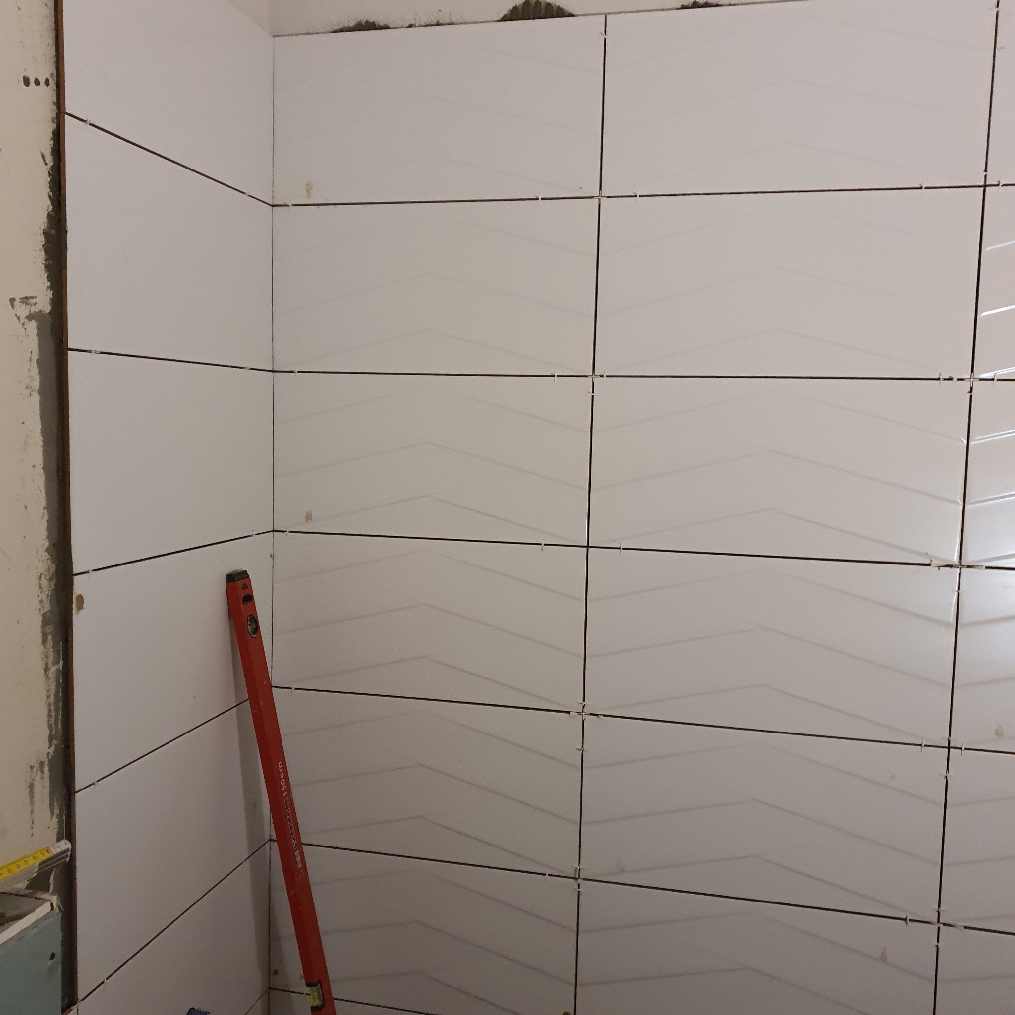 Świeżo ułożone białe płytki ścienne z delikatnym wzorem chevron, poziomowane czerwoną poziomicą, widoczne krzyżyki dystansowe i surowa ściana w tle.