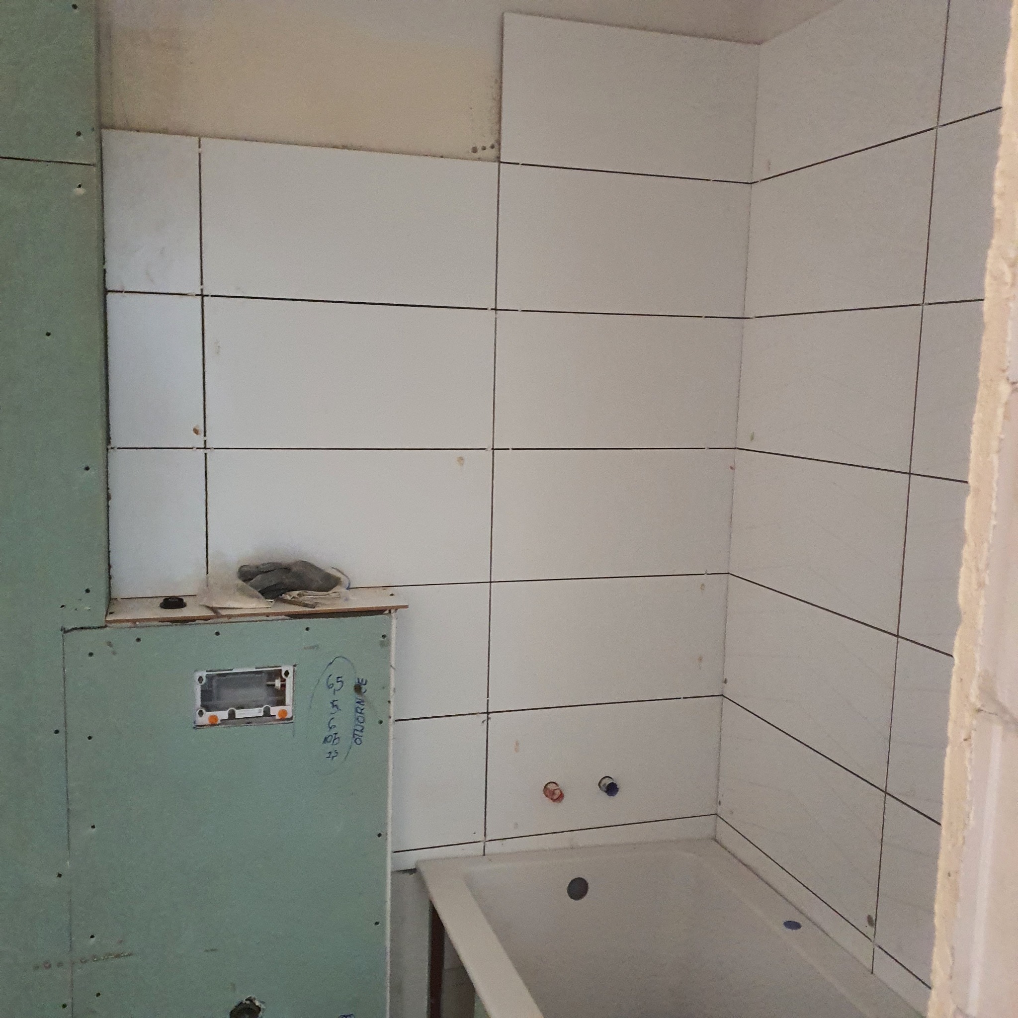 Wnętrze łazienki w trakcie układania białych płytek na ścianach, widoczna wanna, płyta kartonowo-gipsowa z instalacją elektryczną i notatkami odręcznymi, oraz robocze rękawice na półce.
