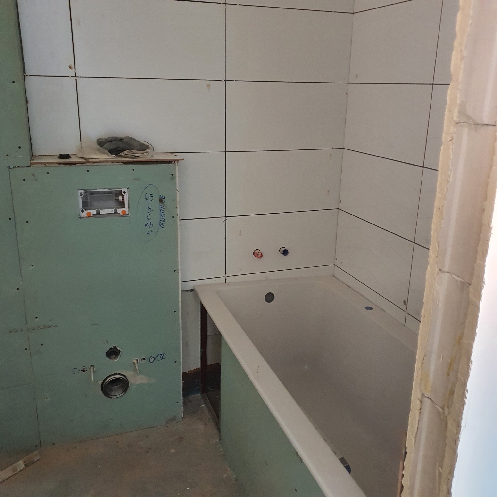 Łazienka w trakcie prac: białe płytki na ścianach, częściowo obudowana wanna, ściana z płyty kartonowo-gipsowej z instalacją hydrauliczną, widoczne niedoskonałości wykończenia przy futrynie.
