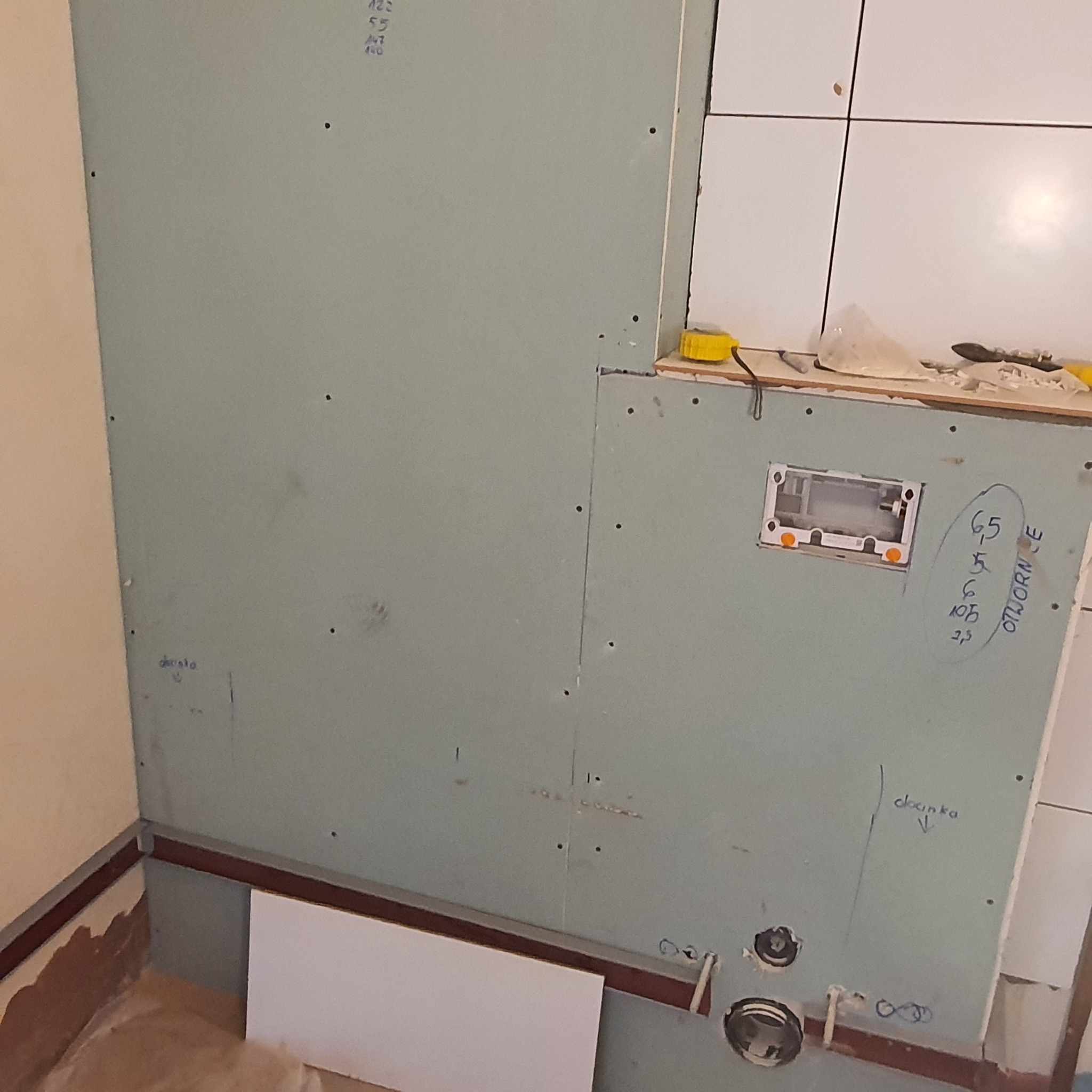 Ściana w trakcie montażu płyt kartonowo-gipsowych w łazience, widoczne otwory na instalacje wodne, szkicowane oznaczenia na płycie oraz fragment ułożonych białych płytek.