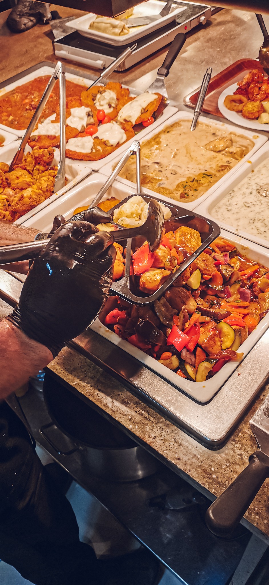 Bufet z różnorodnymi daniami: pieczone warzywa z sosem, kotlety z sosem i pomidorami, smażony kurczak, danie mięsne w sosie, pierścienie cebulowe, całość oświetlona ciepłym światłem.